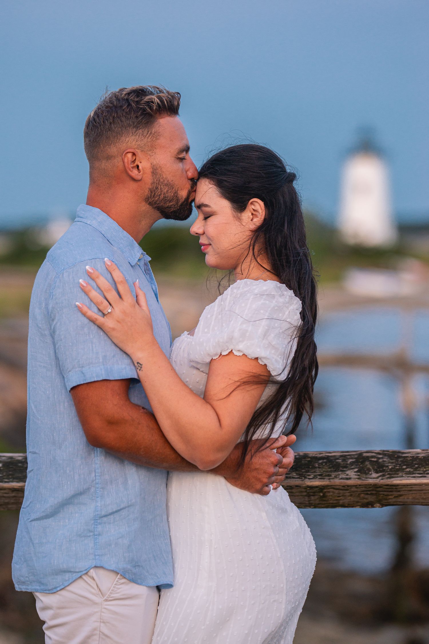 Couple embracing on a waterfront dock, man kissing woman's forehead, lighthouse in background at dusk.