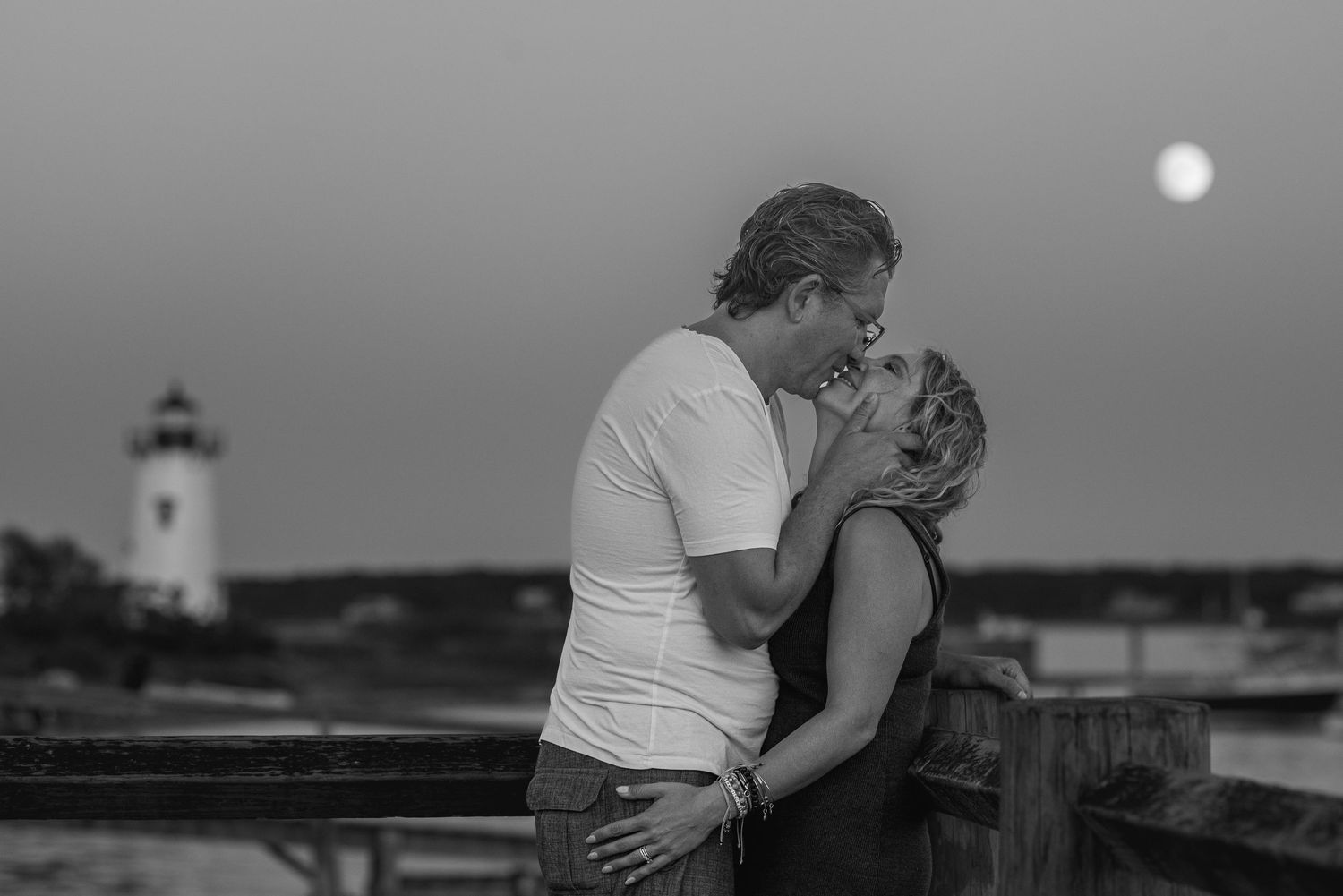 Couple sharing a romantic kiss near a lighthouse under a full moon in a black and white photo.
