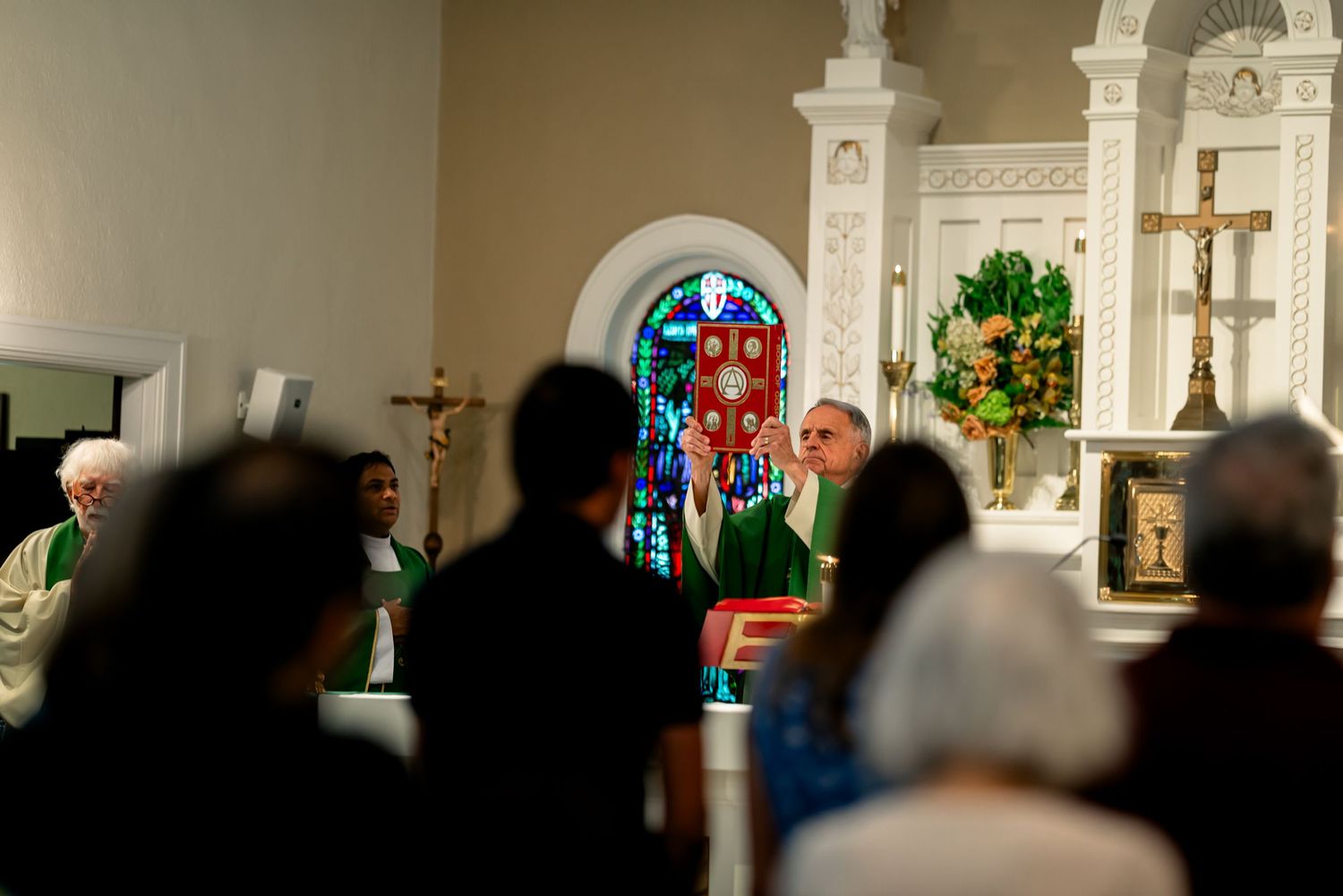 Catholic Mass ceremony inside a church with congregation, priest at altar, stained glass windows, and crucifix.