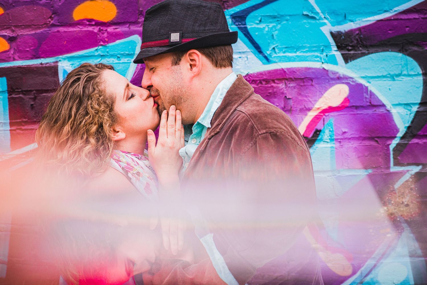 Romantic couple kissing in front of colorful graffiti wall with vibrant pink and blue tones.