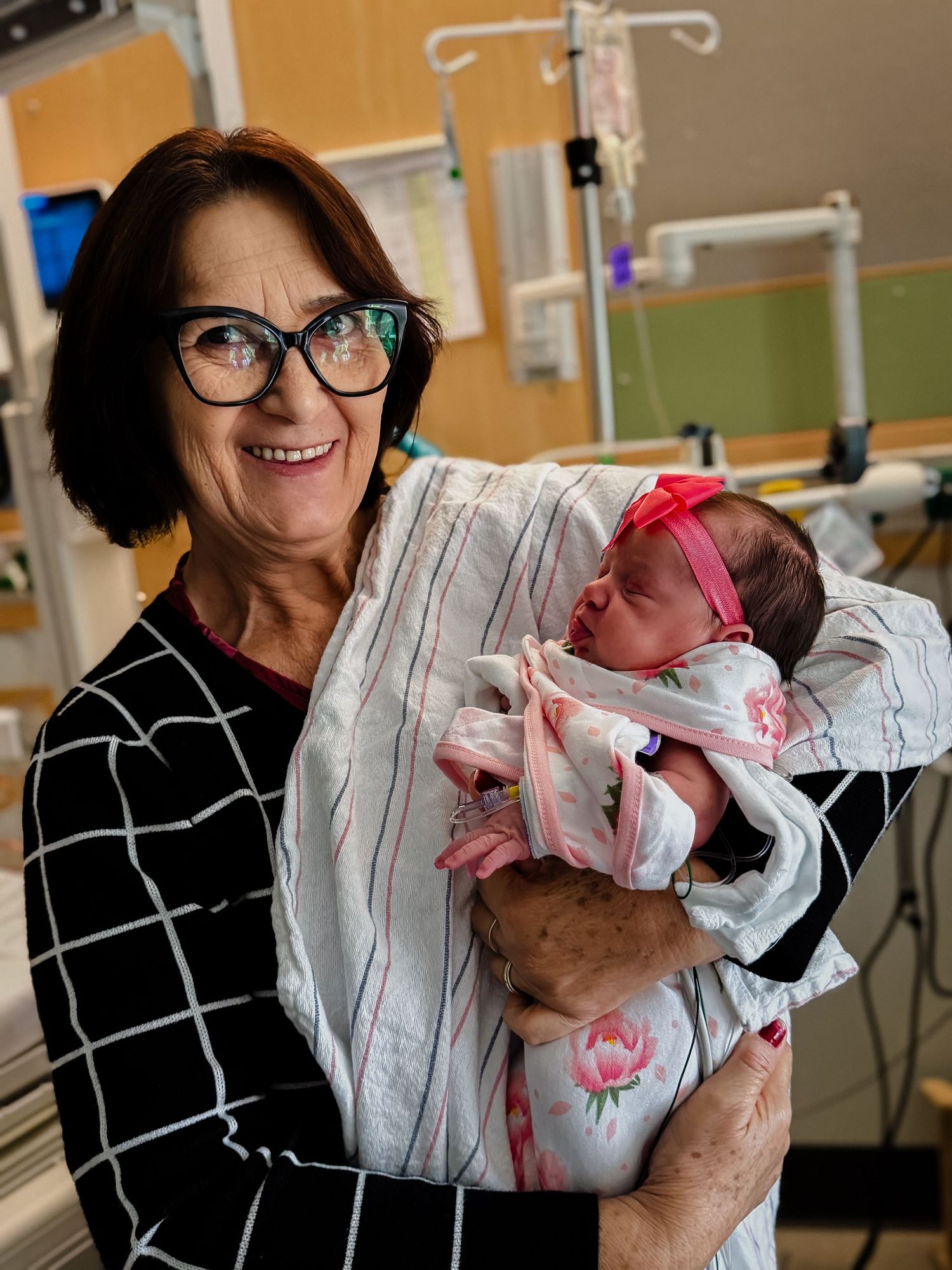 Smiling woman with glasses holds a sleeping newborn in a pink headband and blanket inside a hospital room.