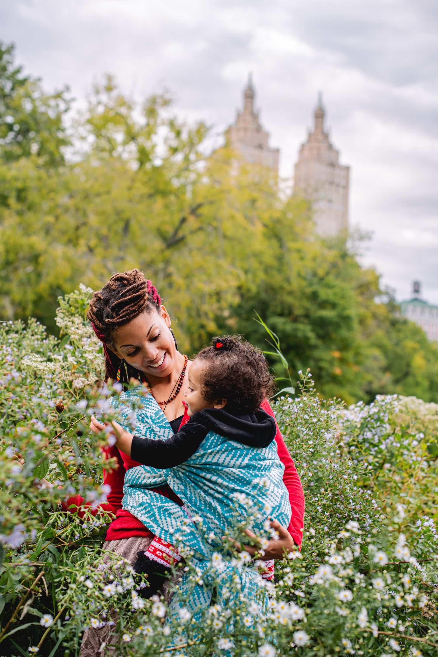 A mother with braided hair holds her baby among white wildflowers in Central Park, NYC, with iconic buildings behind.