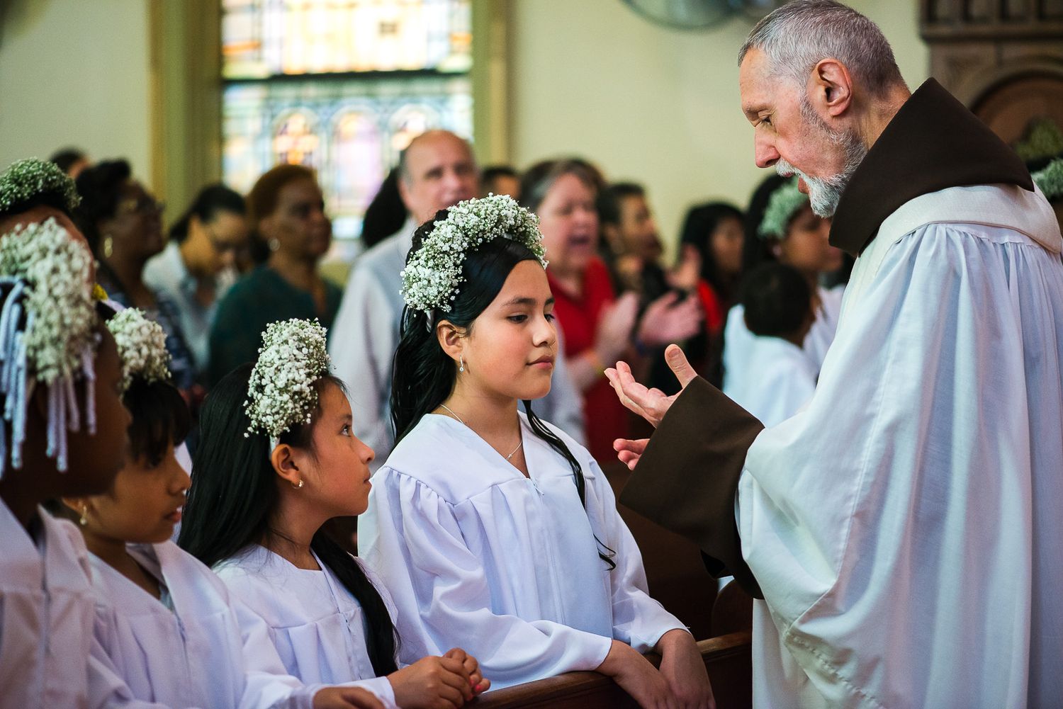 Priest speaking to young girls in white dresses with floral headpieces during a First Communion ceremony in a church.