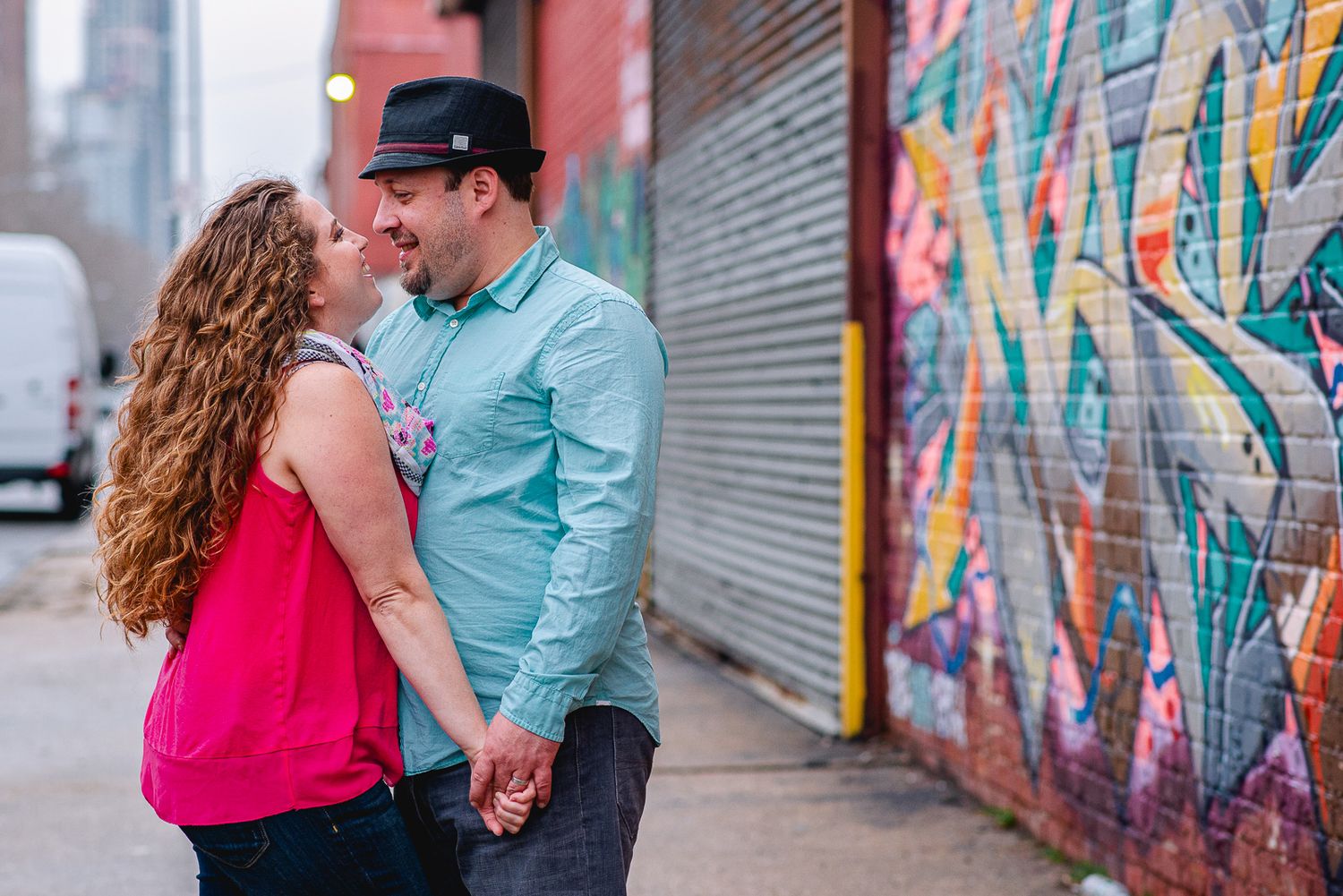 Couple sharing a romantic kiss while holding hands in front of a colorful graffiti wall on a city sidewalk.