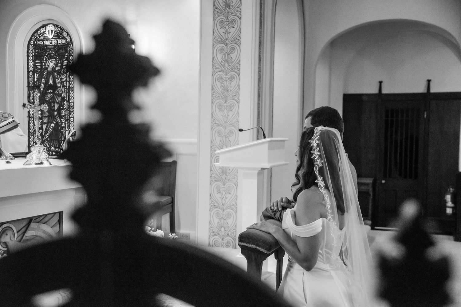 Bride in white wedding gown and veil kneeling at church altar during wedding ceremony, black and white photo.