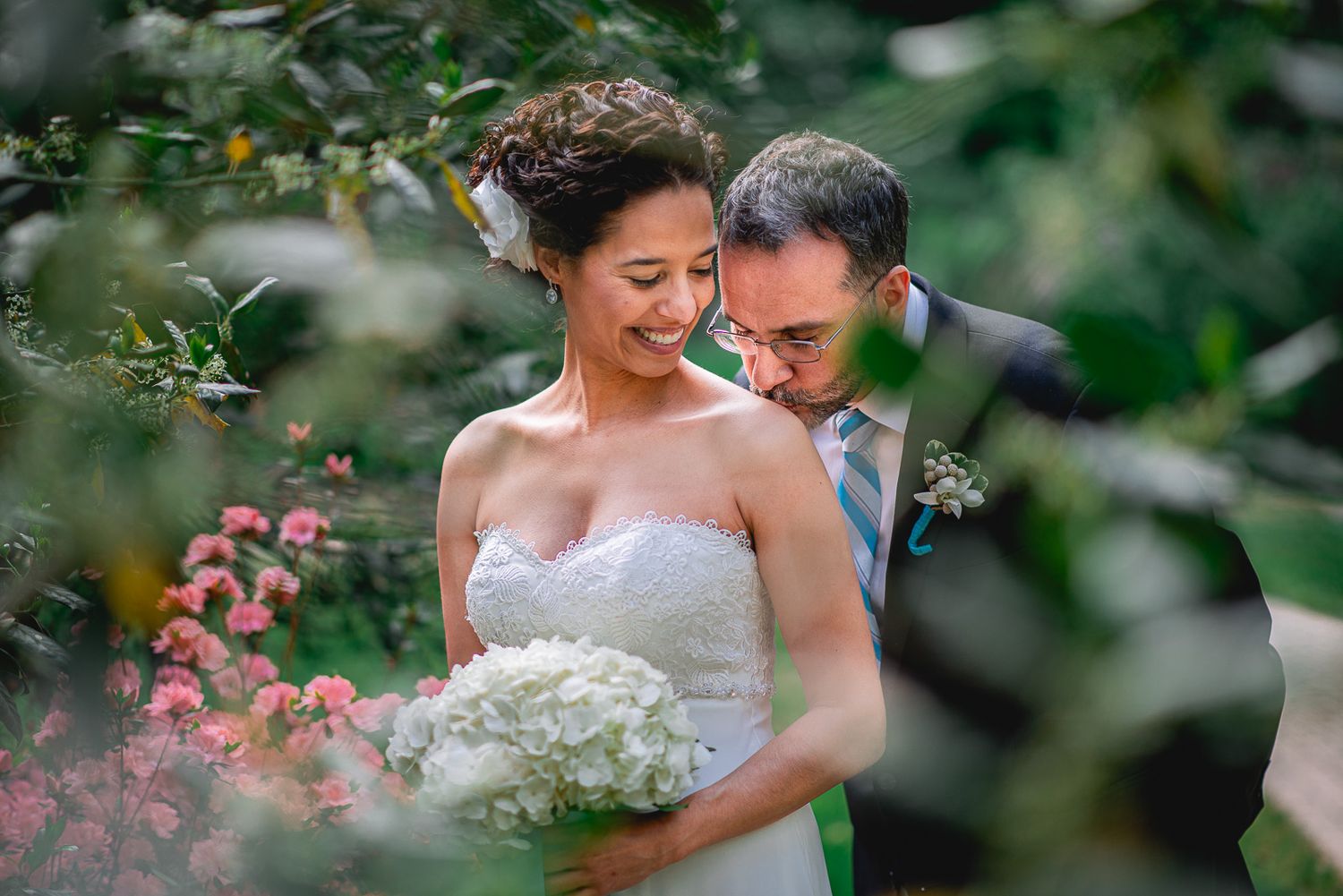 Bride and groom share a tender moment surrounded by lush greenery and pink flowers, bride holding white hydrangea bouquet.