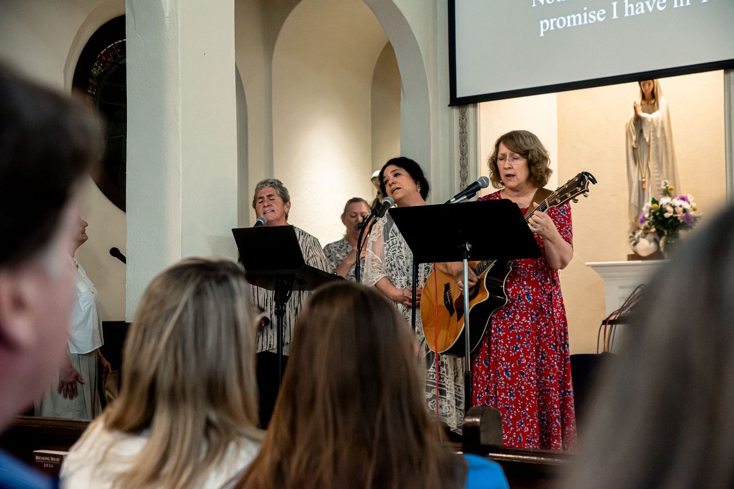 Women singing and playing guitar during a church worship service with a projection screen displaying lyrics.