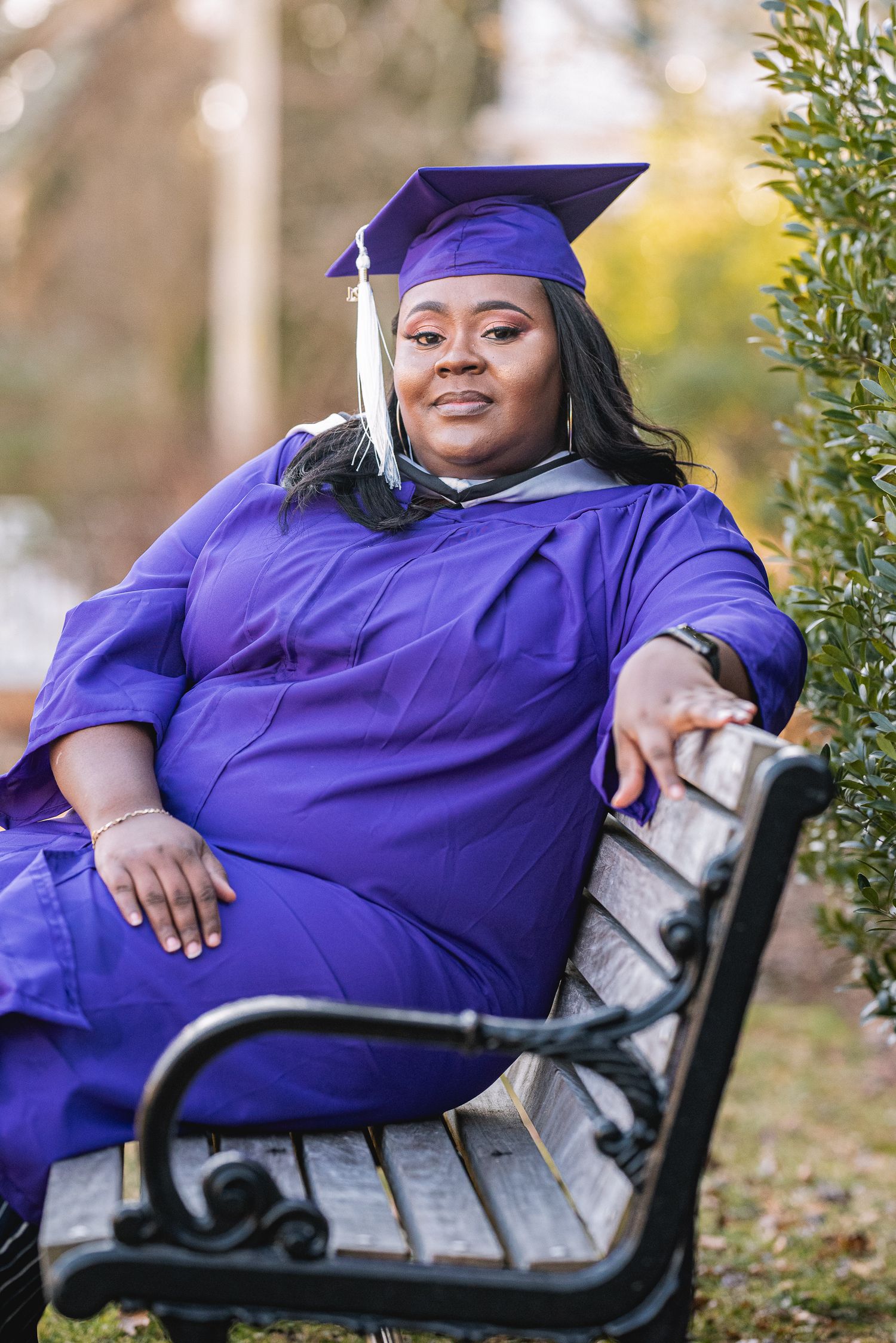 A woman in a purple graduation cap and gown sits relaxed on a park bench, smiling proudly outdoors.