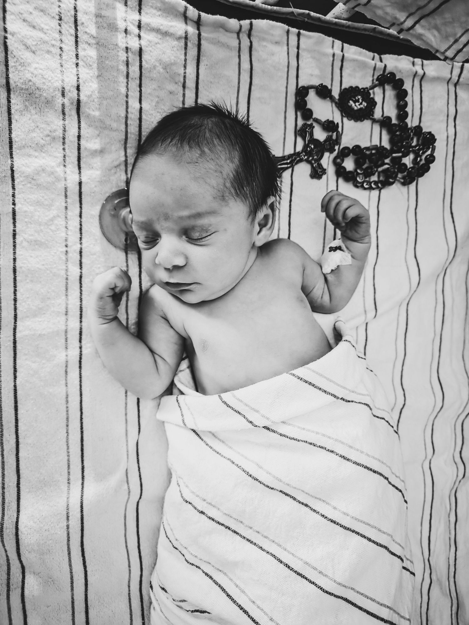 Black and white photo of a sleeping newborn baby swaddled in a striped blanket, with a pacifier and a rosary resting beside the infant.