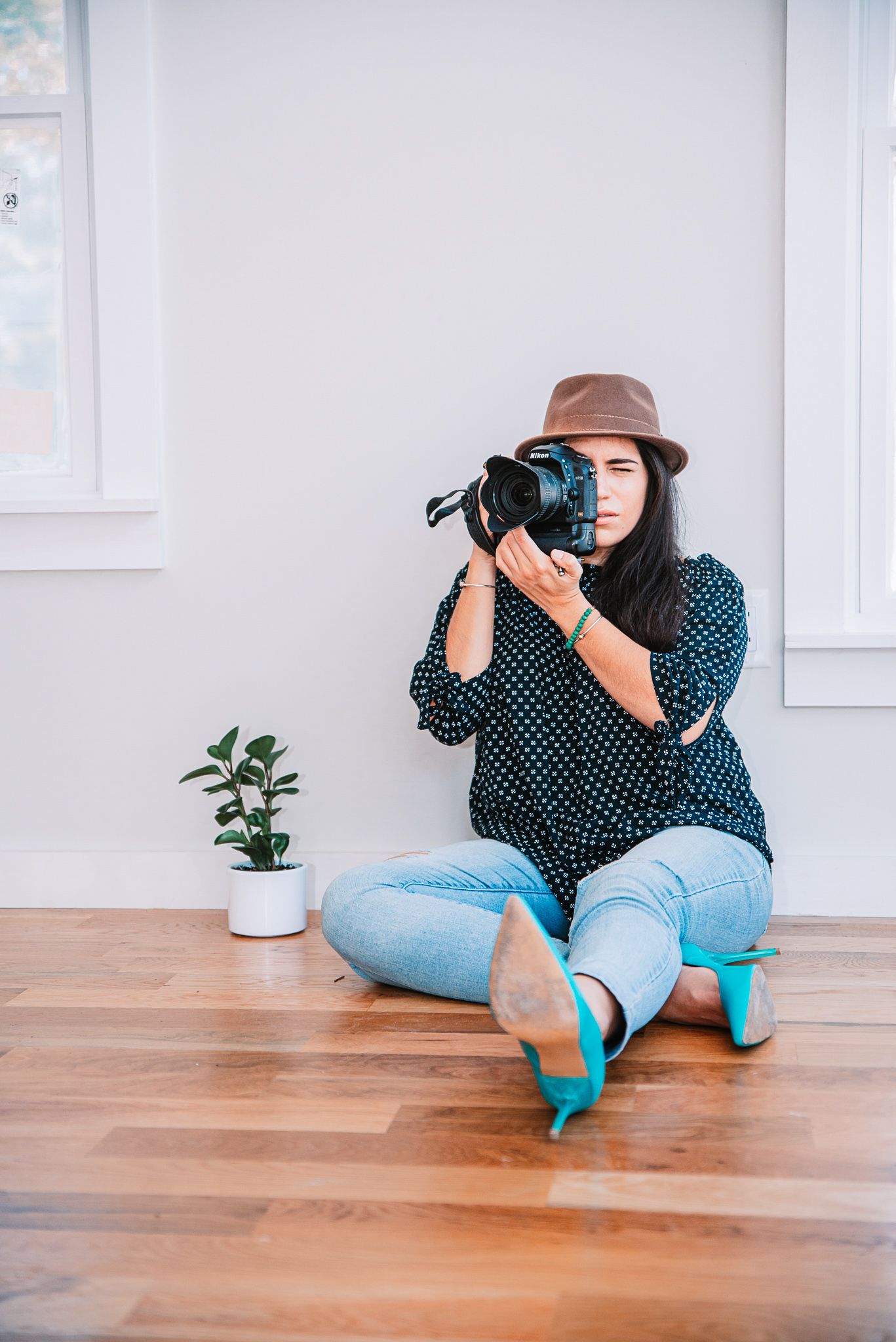Female photographer sitting on floor aiming DSLR camera, wearing hat and teal heels.