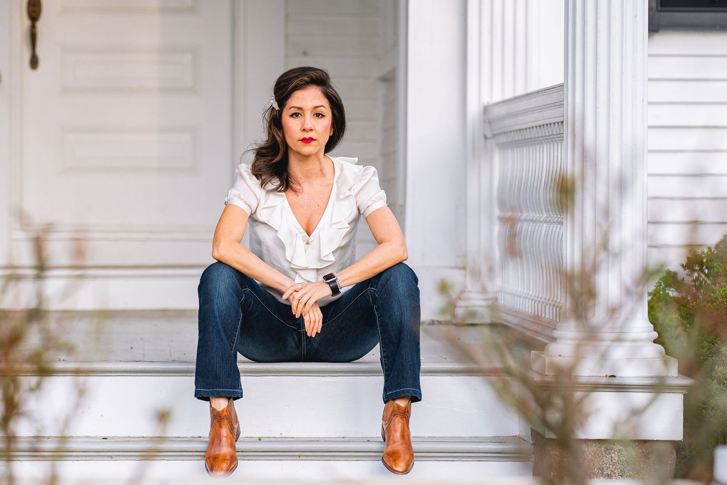 Woman in white ruffled blouse and jeans sits on white porch steps wearing brown boots.