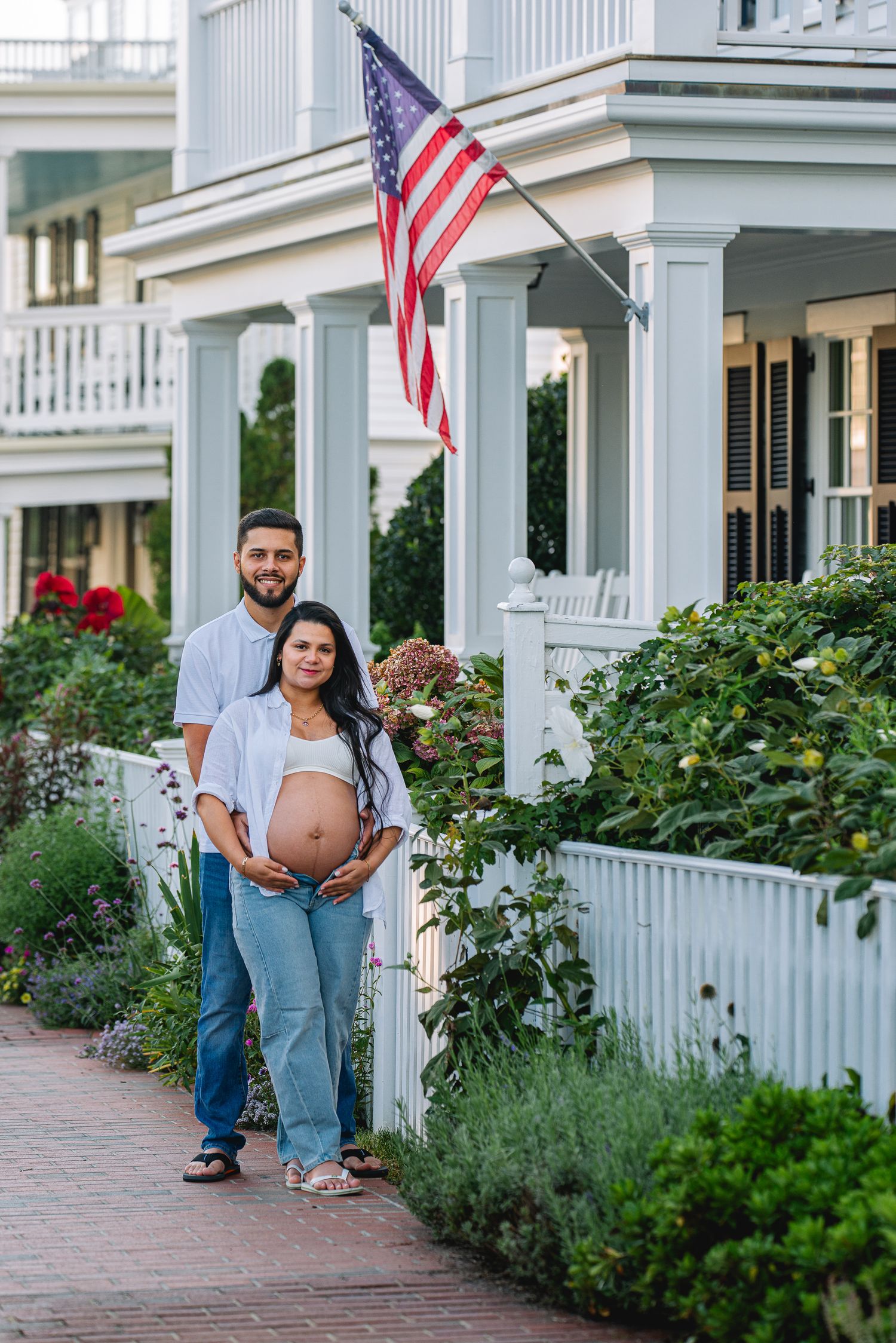 Pregnant couple poses outside a white colonial home with an American flag, surrounded by lush gardens on a brick pathway.