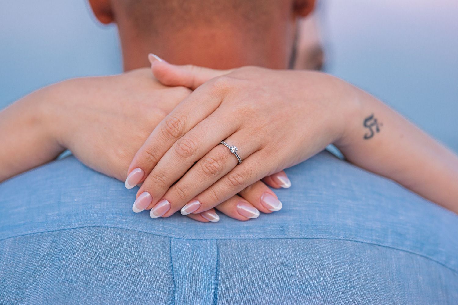 Close-up of a woman's hand with a diamond engagement ring resting on a man's back, with a tattoo visible.