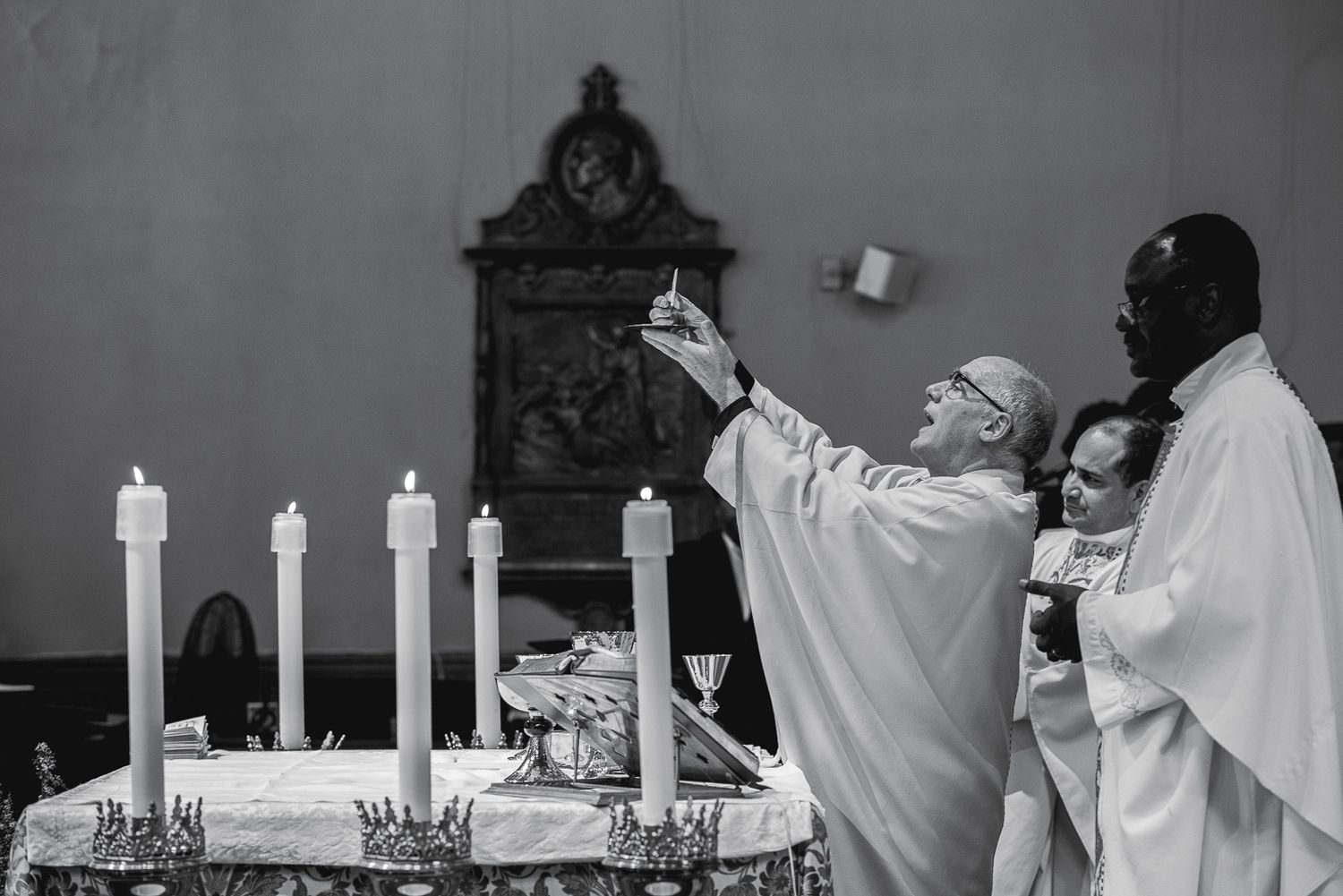 Black and white photo of priests performing a Catholic Mass ceremony at an altar with lit candles in a church.