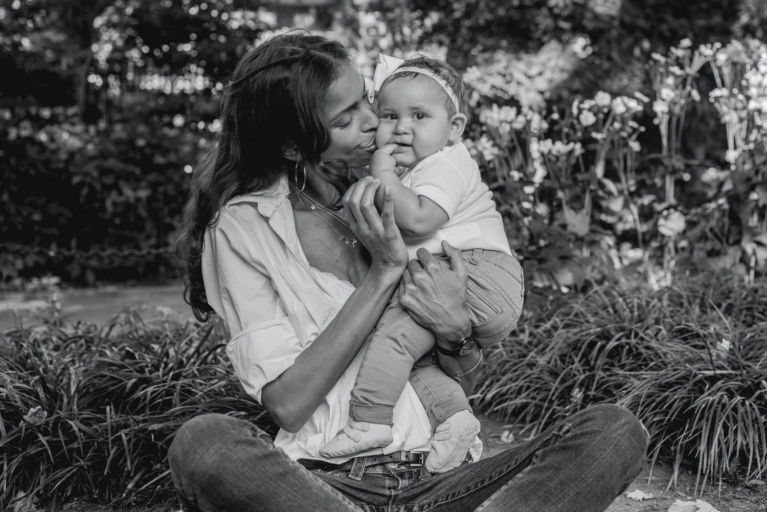 Black and white photo of a mother kissing her baby while sitting in a lush garden.