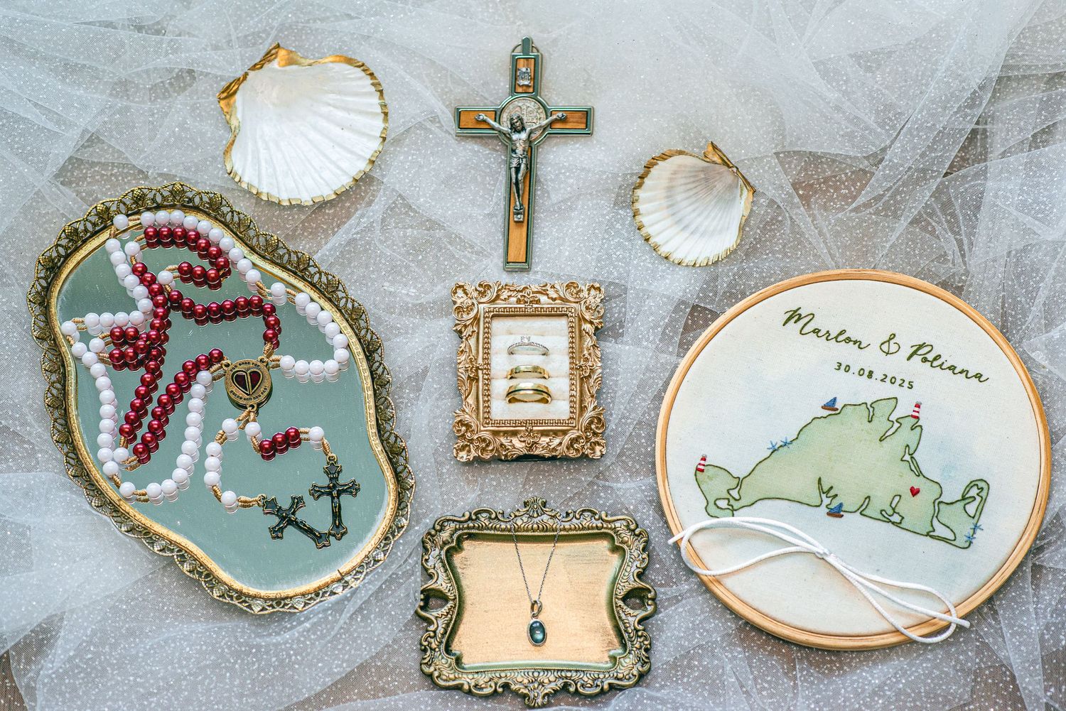 Wedding flat lay with rosary beads, crucifix, embroidery hoop, framed rings, necklace, and seashells on white tulle.