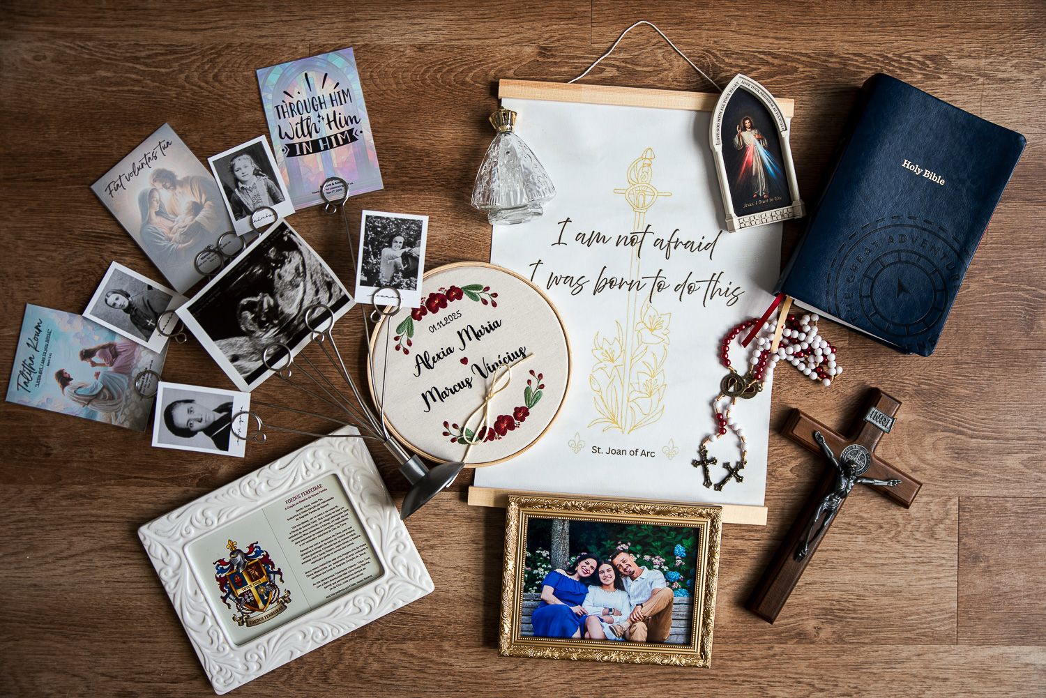Flat lay of Catholic keepsakes on wood with photos, rosary, crucifix, embroidered hoop, prayer card, bible and a framed family picture.