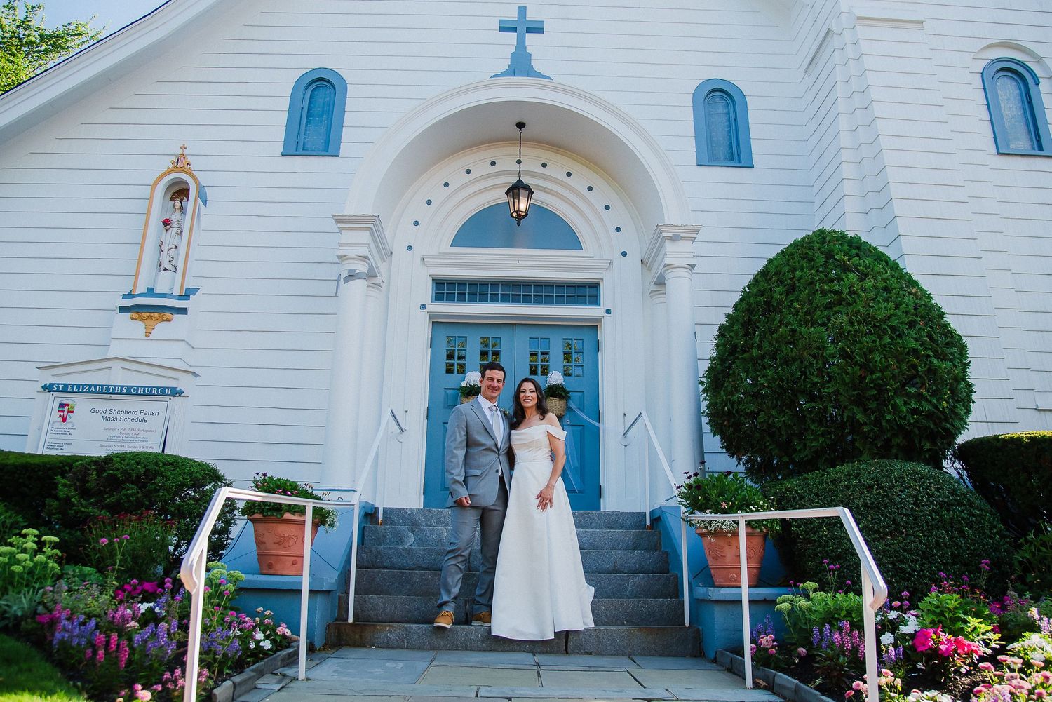 Bride and groom pose on steps of white church with blue doors and cross, surrounded by lush greenery and flowers.