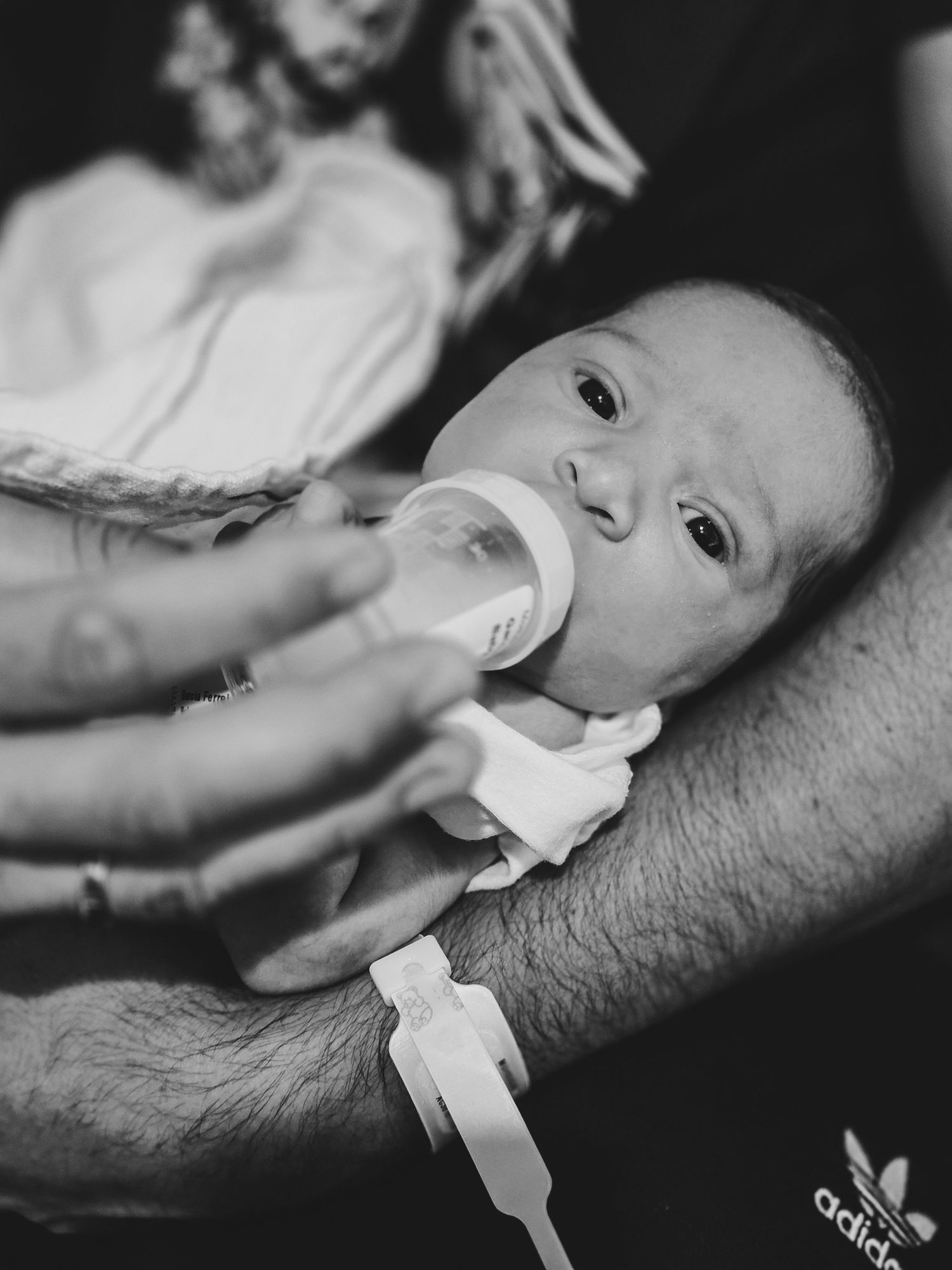Black and white photo of a newborn baby being bottle-fed while cradled in an adult's tattooed arm, with a hospital ID bracelet.