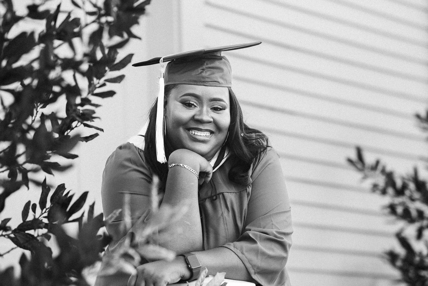 Smiling graduate in cap and gown posing outdoors among green leaves in a black and white photo.