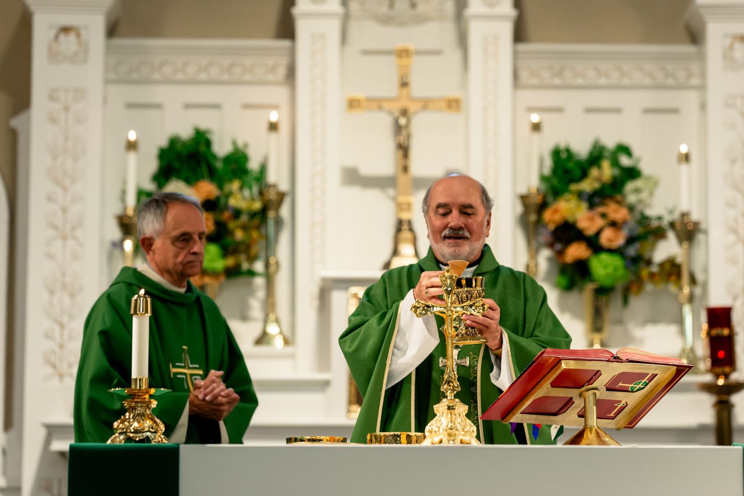 Two Catholic priests in green vestments celebrate Mass at an ornate white altar with candles and floral arrangements.