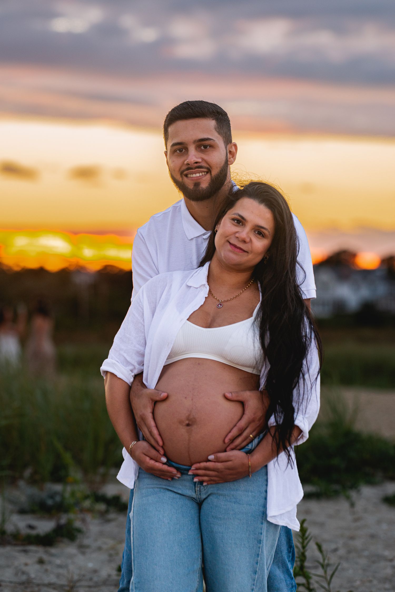 Happy couple embracing at sunset, pregnant woman in white crop top and jeans cradling baby bump outdoors.