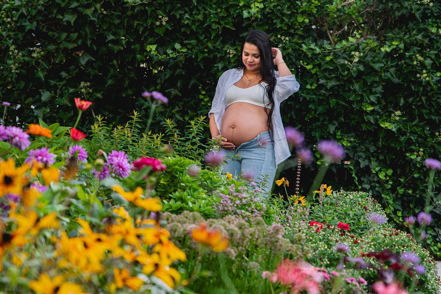 Pregnant woman in a colorful flower garden, wearing jeans and a crop top, cradling her baby bump outdoors.
