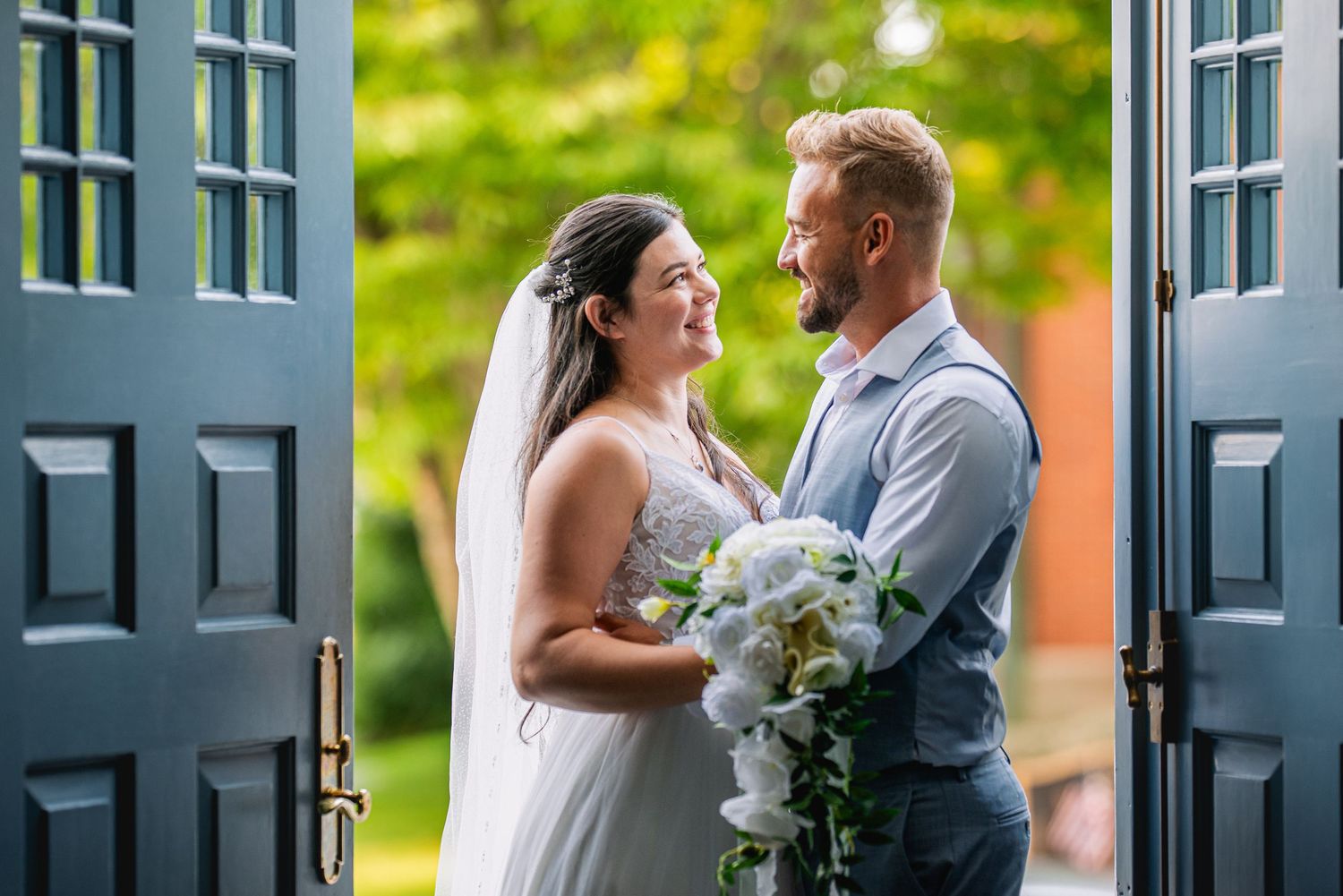 Bride and groom share a romantic moment in a church doorway, bride holding white flowers, lush greenery in background.