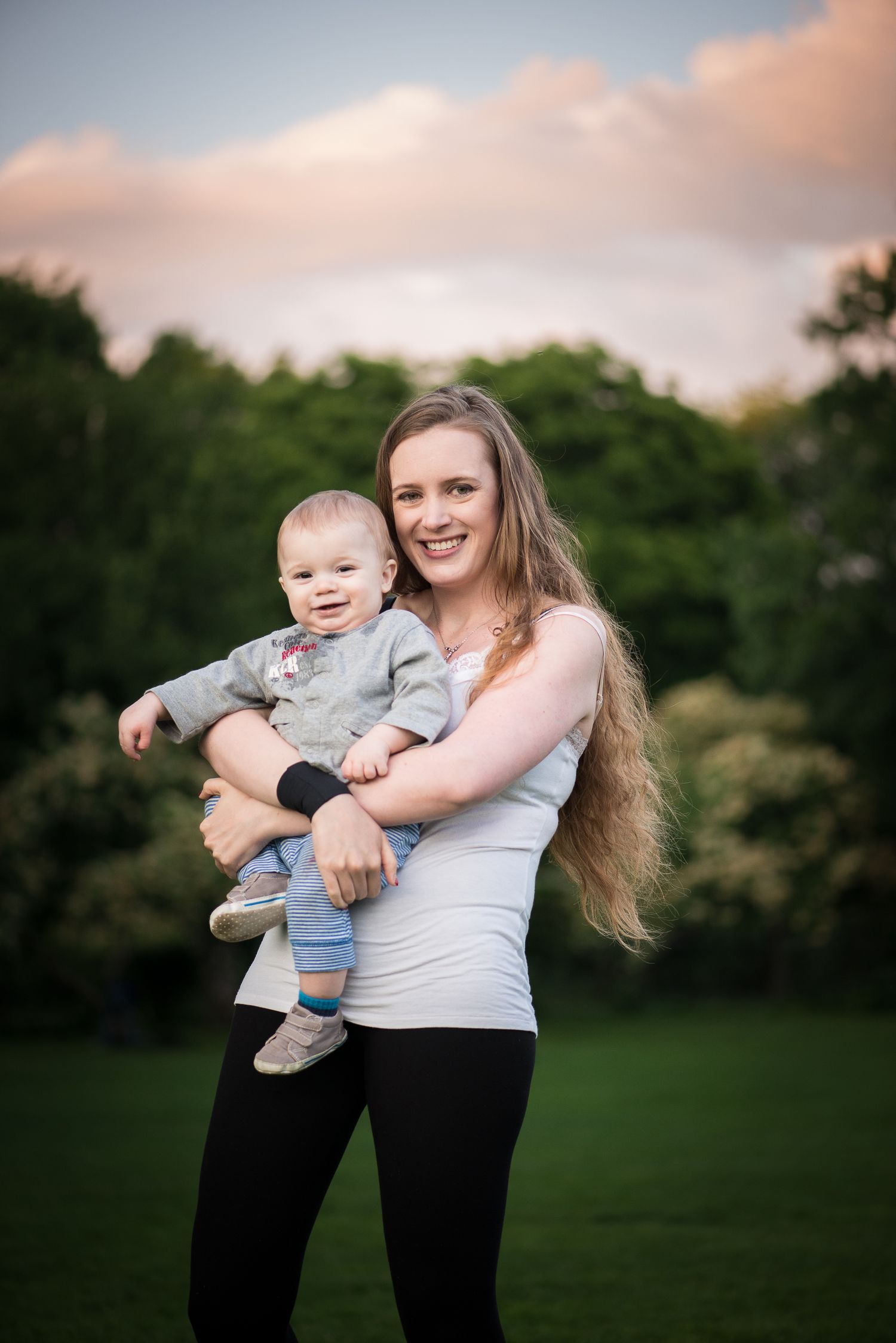 Smiling mother with long blonde hair holding baby boy in park at golden hour sunset.