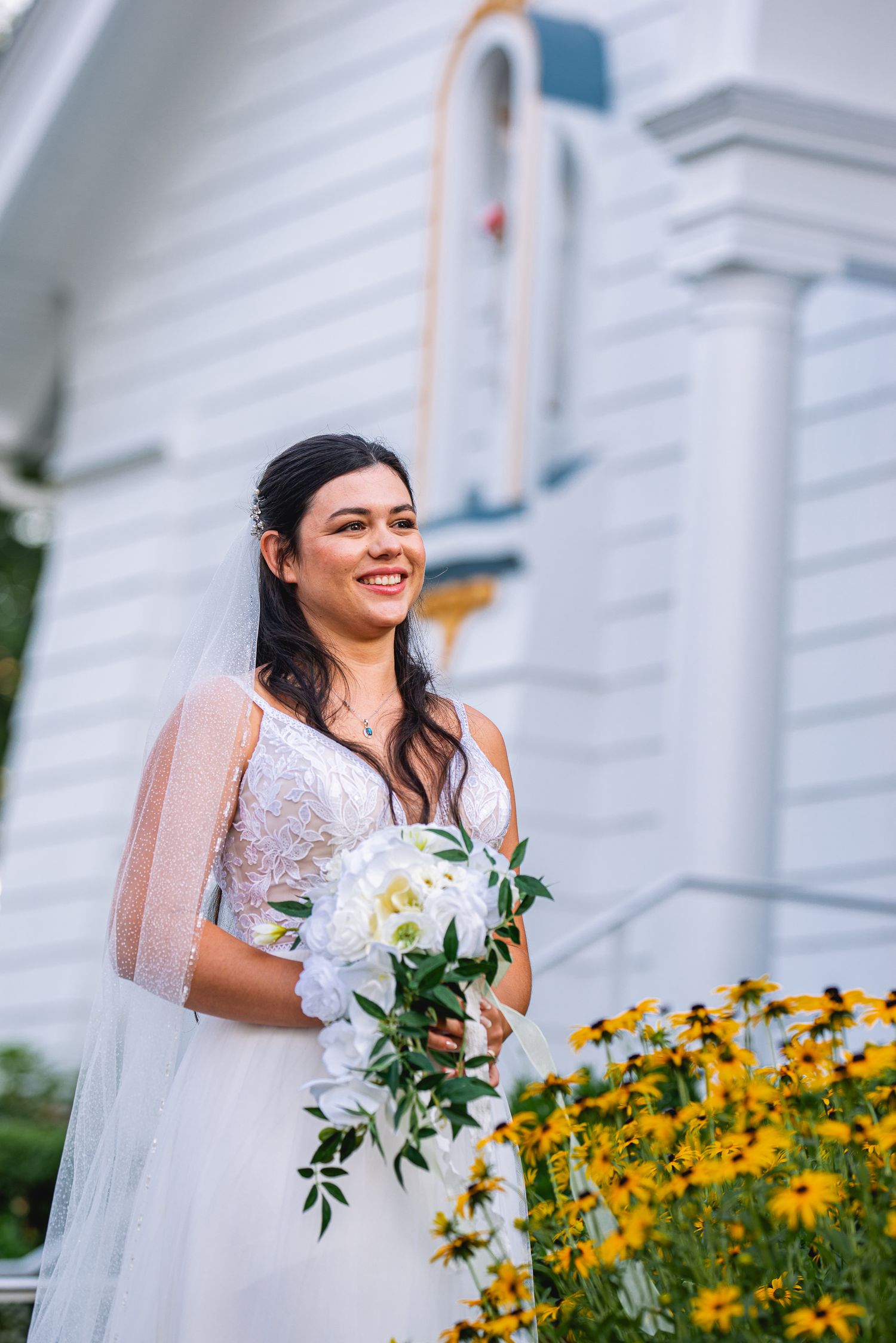 Smiling bride in lace wedding gown holding white bouquet stands beside yellow flowers outside white church.
