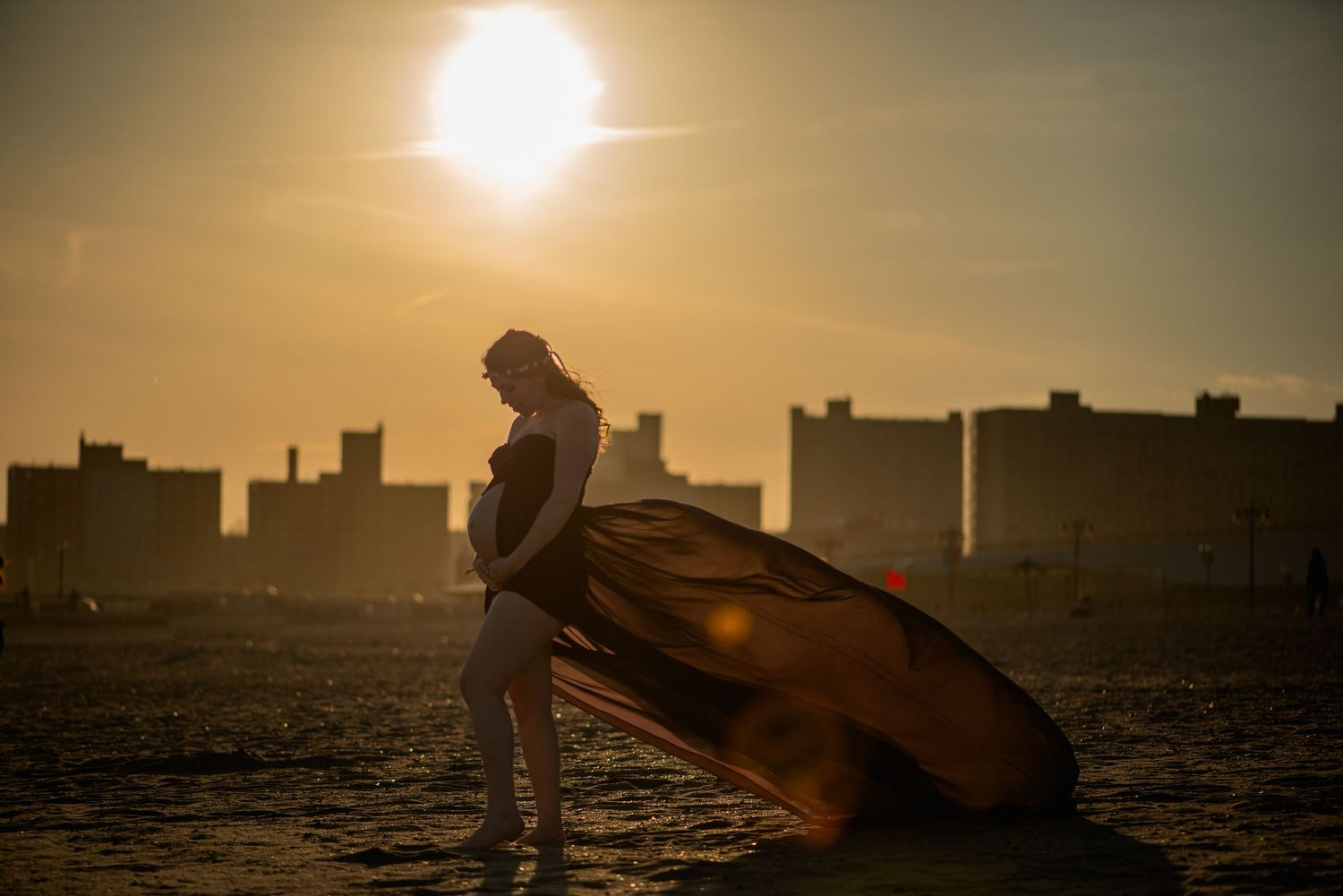 Pregnant woman in white dress with flowing golden fabric at sunset on beach with city skyline silhouette.