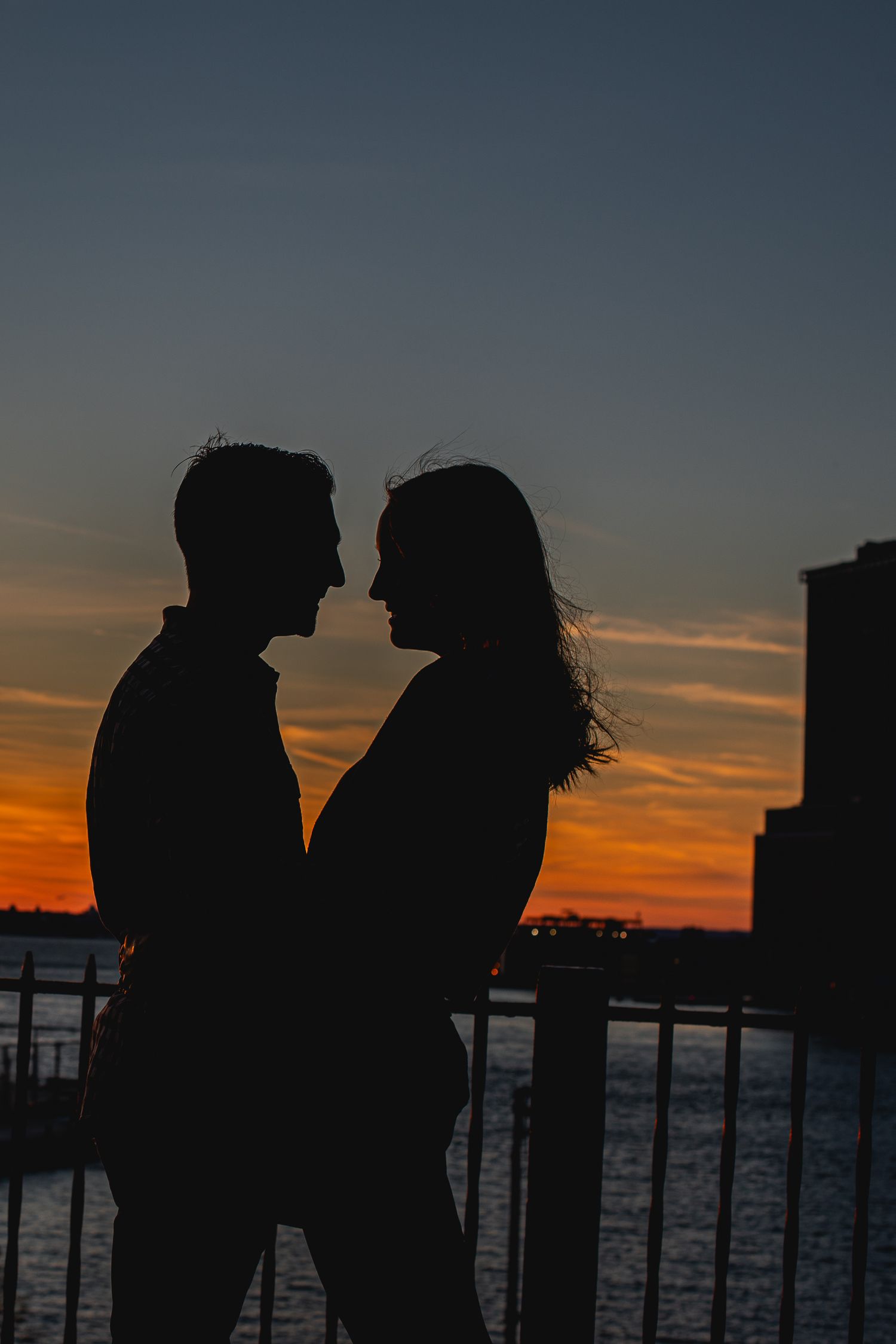 Romantic silhouette of a couple facing each other against a vibrant orange and blue sunset sky near a waterfront.