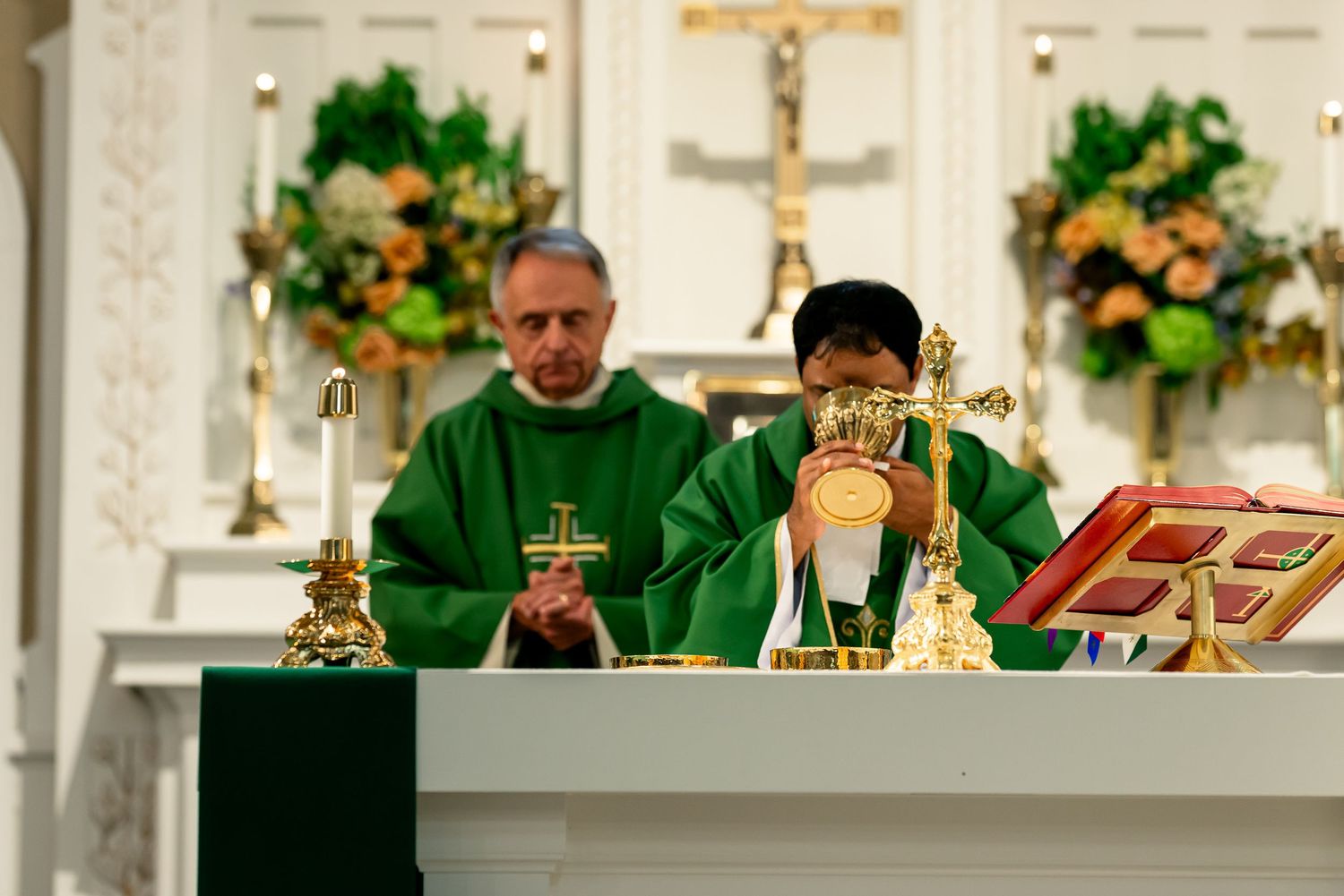 Two Catholic priests celebrate Mass at an altar in a white church, wearing green vestments during liturgy.