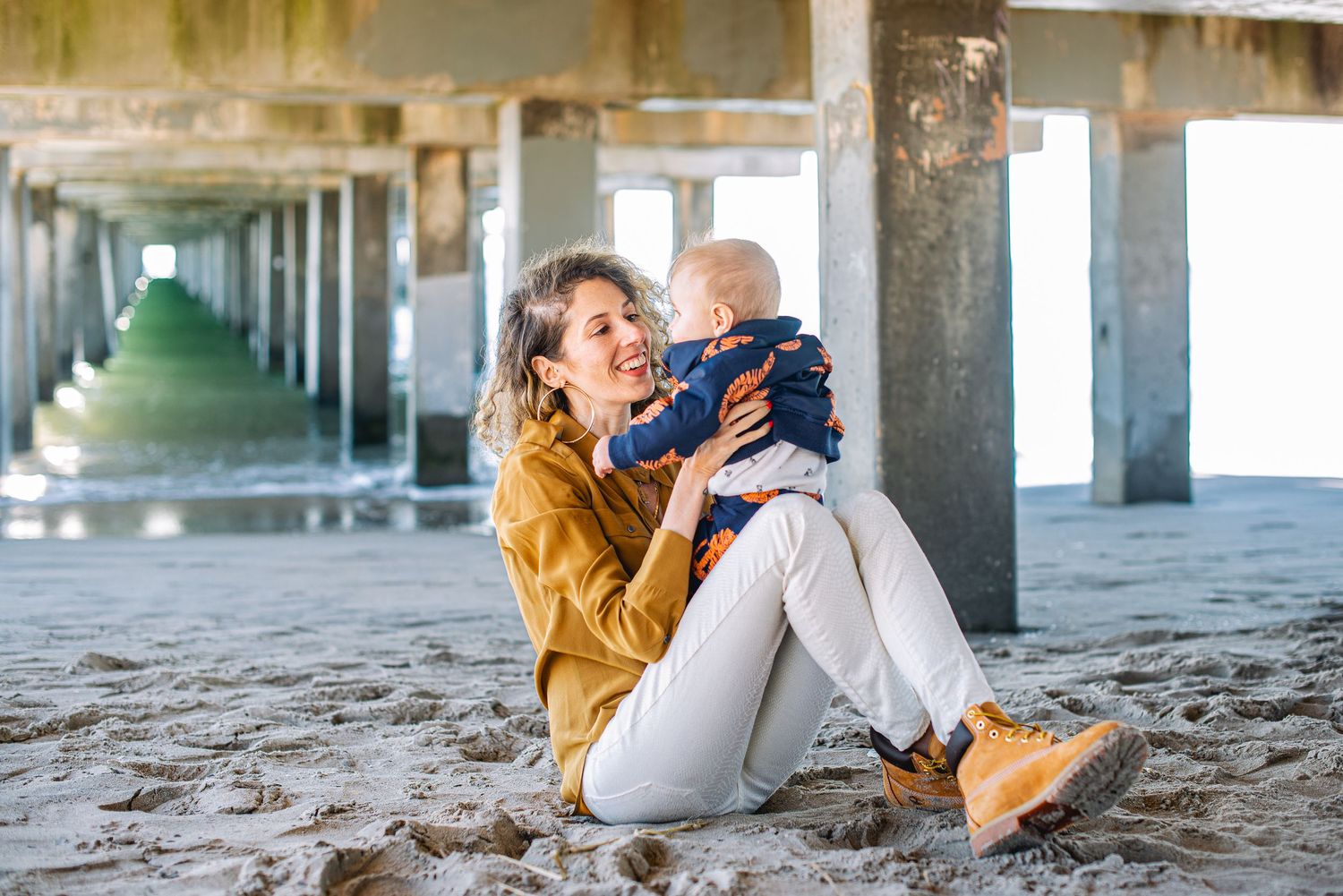 A mother sits on sandy beach under a pier, lovingly holding her baby in the air on a sunny day.