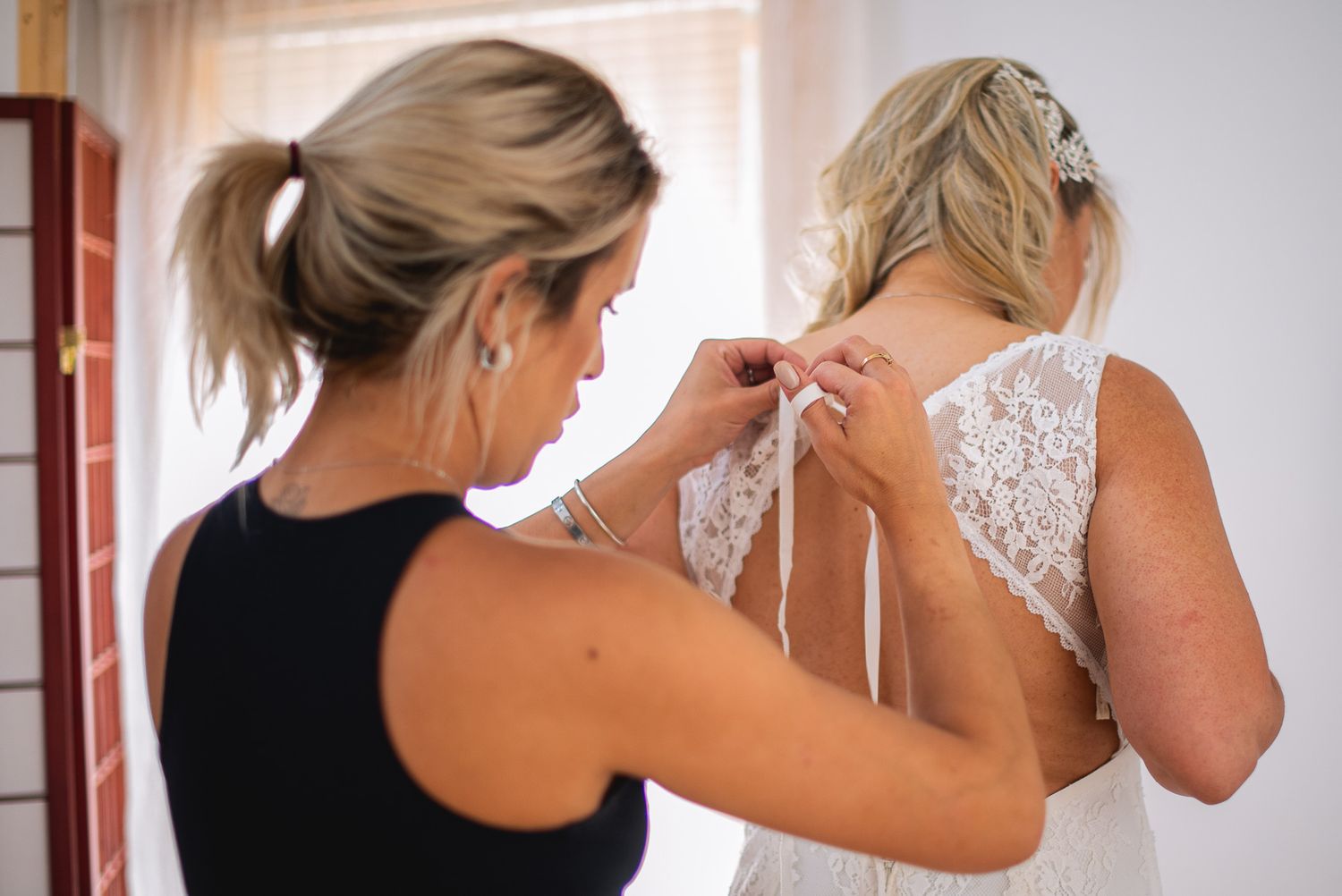 Bridesmaid helping bride lace up back of white wedding dress before ceremony.