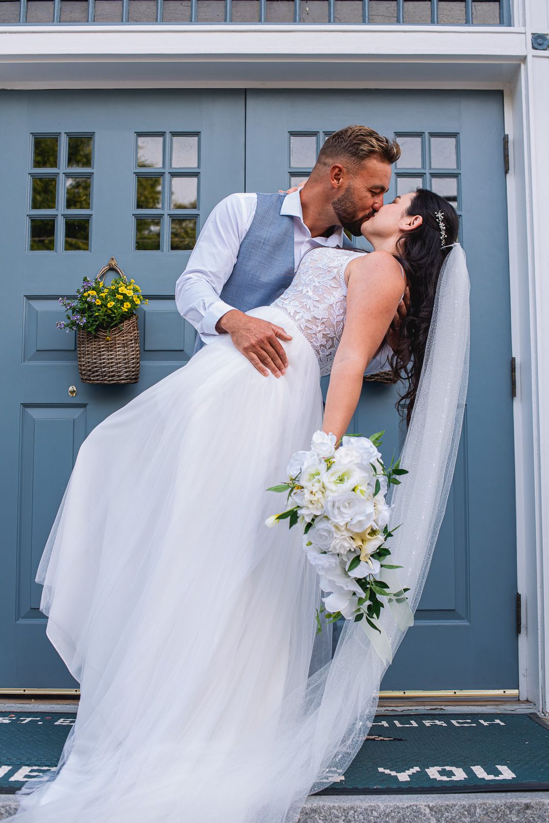 Bride and groom sharing a romantic dip kiss in front of blue church doors with white bouquet.