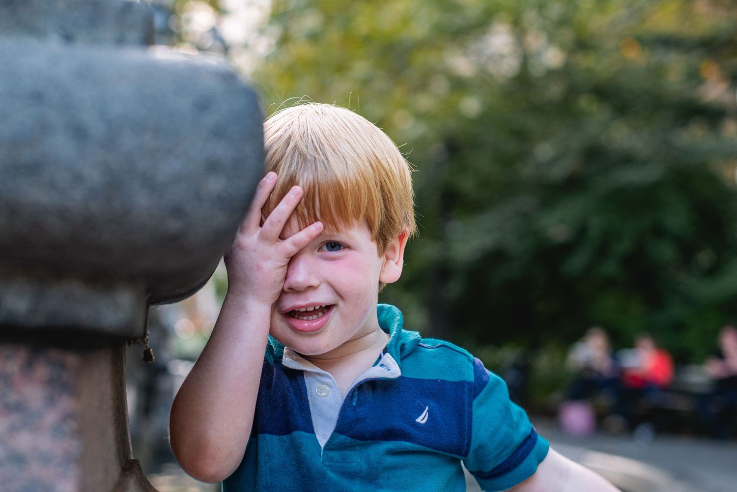 Smiling blonde boy covering one eye with hand near stone fountain in outdoor park setting.