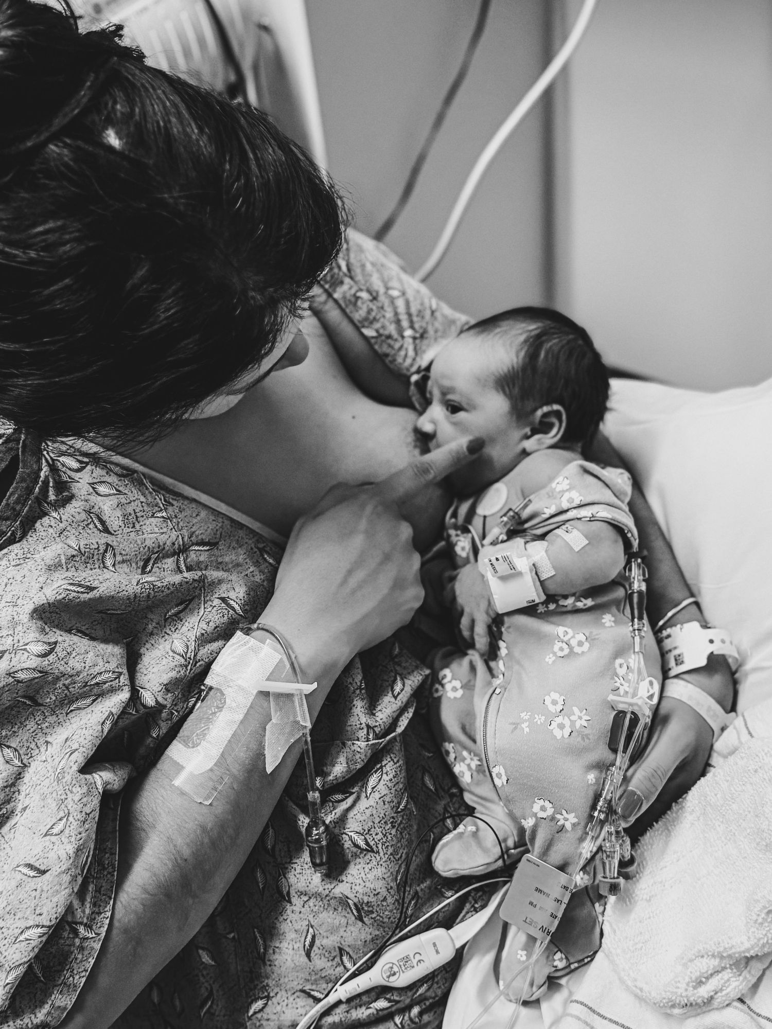 Black and white photo of a new mother in a hospital gown with an IV bracelet gently holding her sleeping newborn baby in a hospital bed.