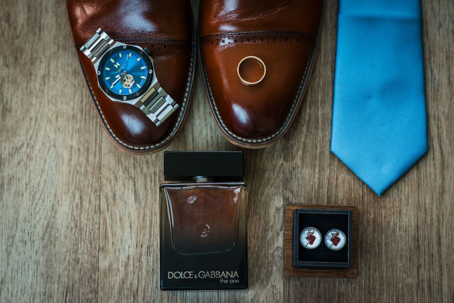 Brown leather dress shoes, blue watch, gold ring, Dolce & Gabbana cologne, blue tie, and cufflinks arranged on wood floor.