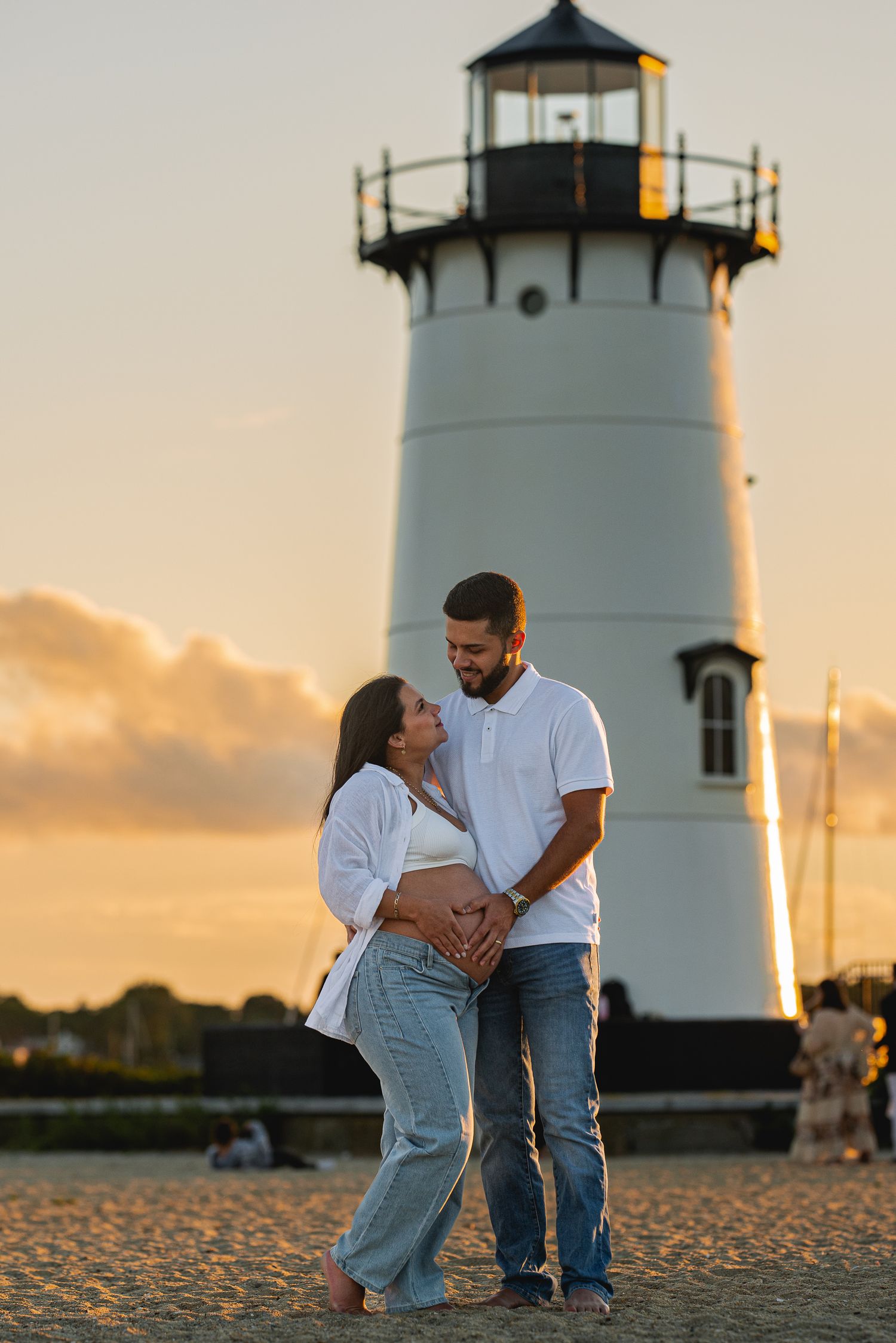 Pregnant couple embracing at sunset in front of a white lighthouse on a sandy beach.