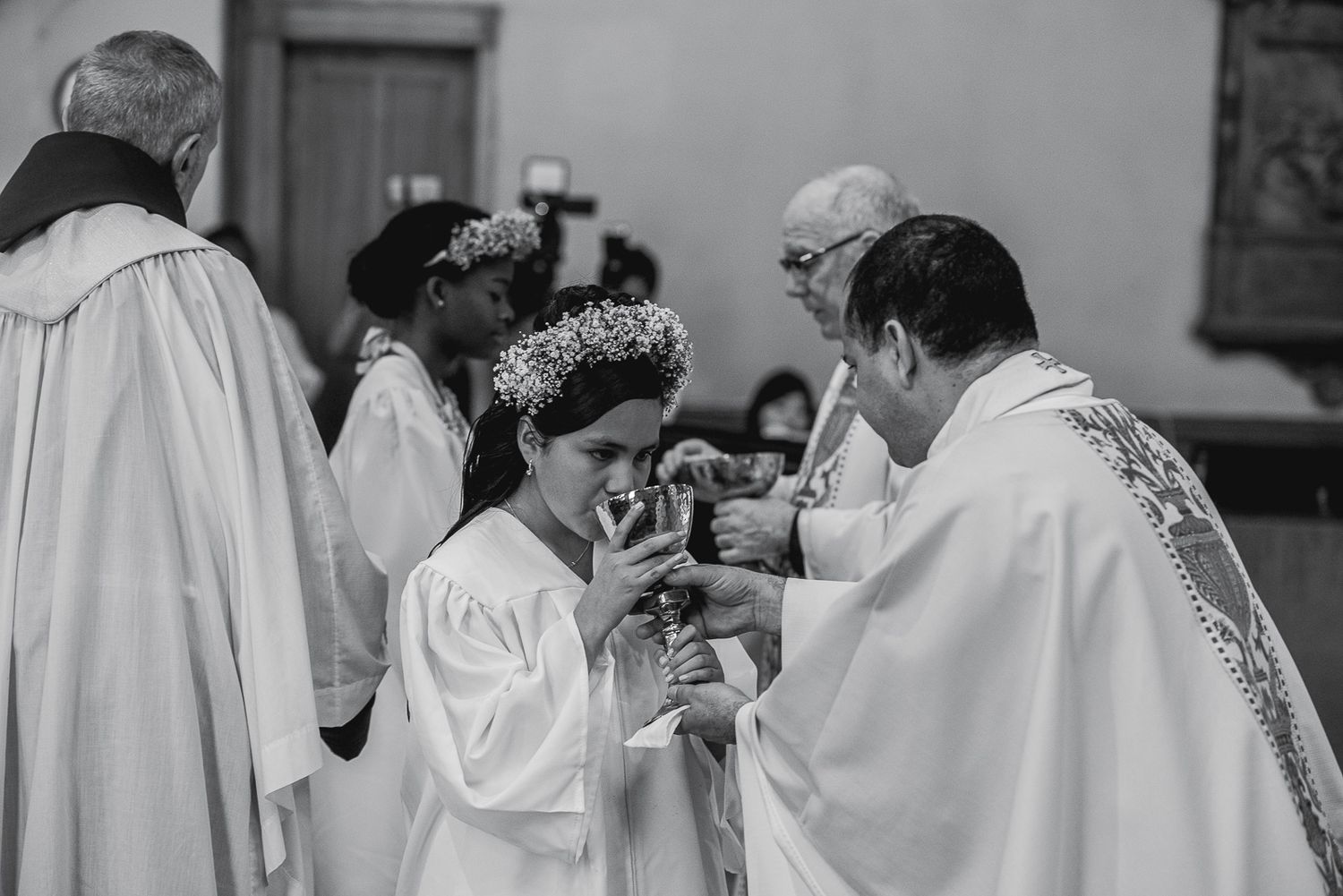 Black and white photo of a young girl in white dress receiving First Communion from a priest in a church.