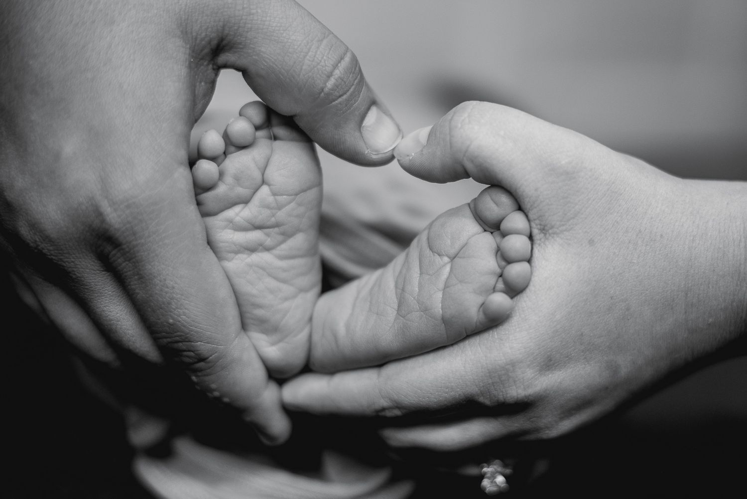 Black and white photo of adult hands cradling tiny newborn baby feet in a heart shape.