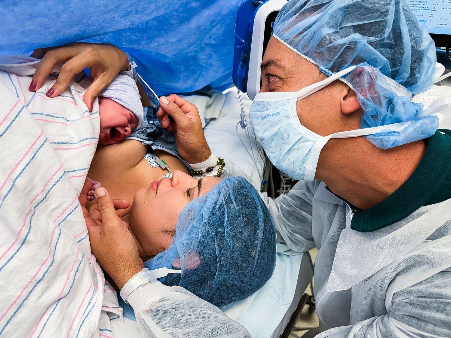 A masked doctor in surgical scrubs helps a mother in a blue cap hold her newborn baby in the operating room after a cesarean delivery.