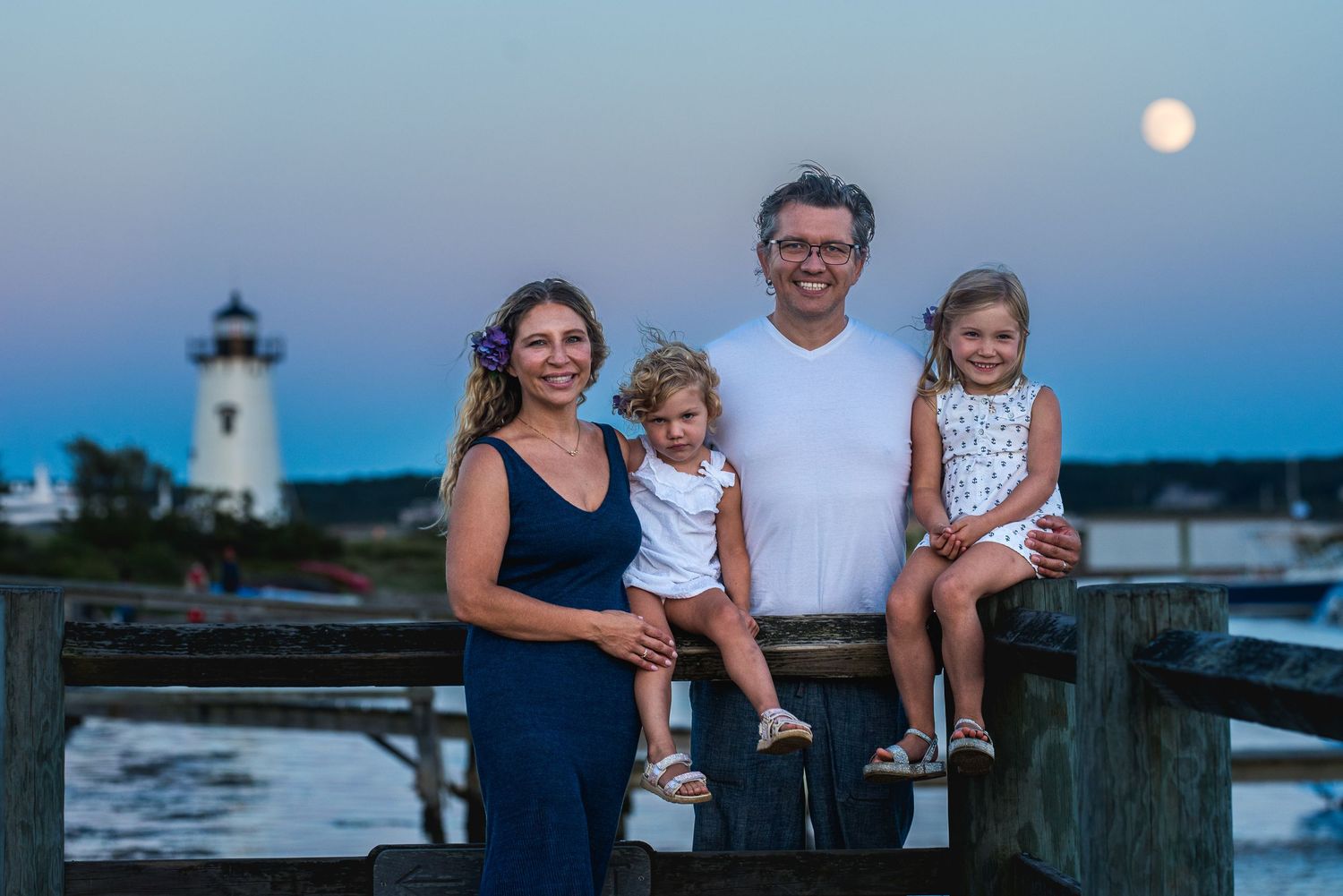 Family of four smiling on a dock at dusk with a lighthouse and full moon in the background.