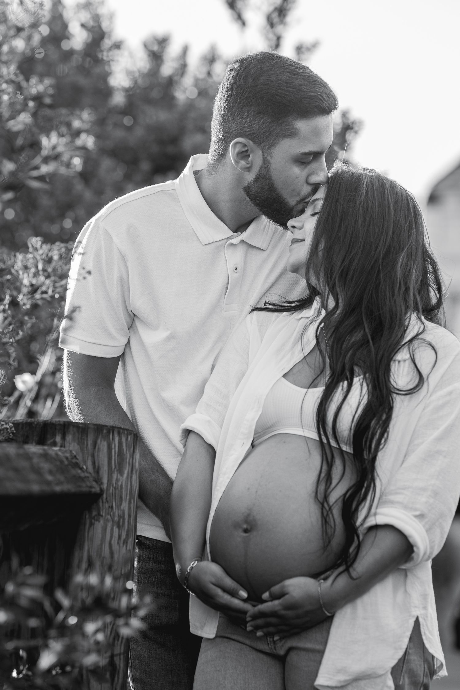 Black and white photo of a man kissing a pregnant woman's forehead while they both cradle her baby bump outdoors.