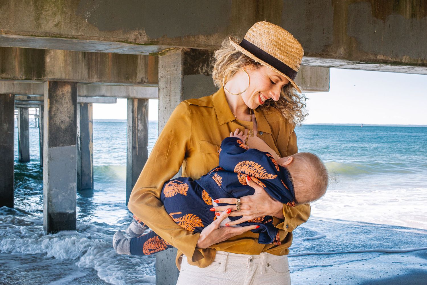 A woman in a straw hat holds a sleeping newborn baby by the ocean under a pier.