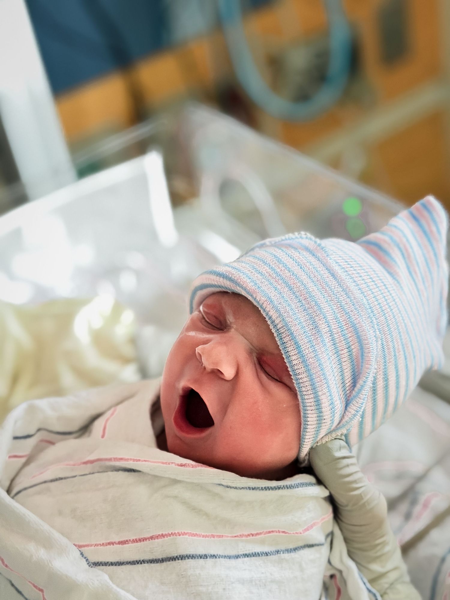 Newborn baby in a striped hospital hat and blanket yawning with mouth wide open while a gloved hand gently supports the head in a bassinet.