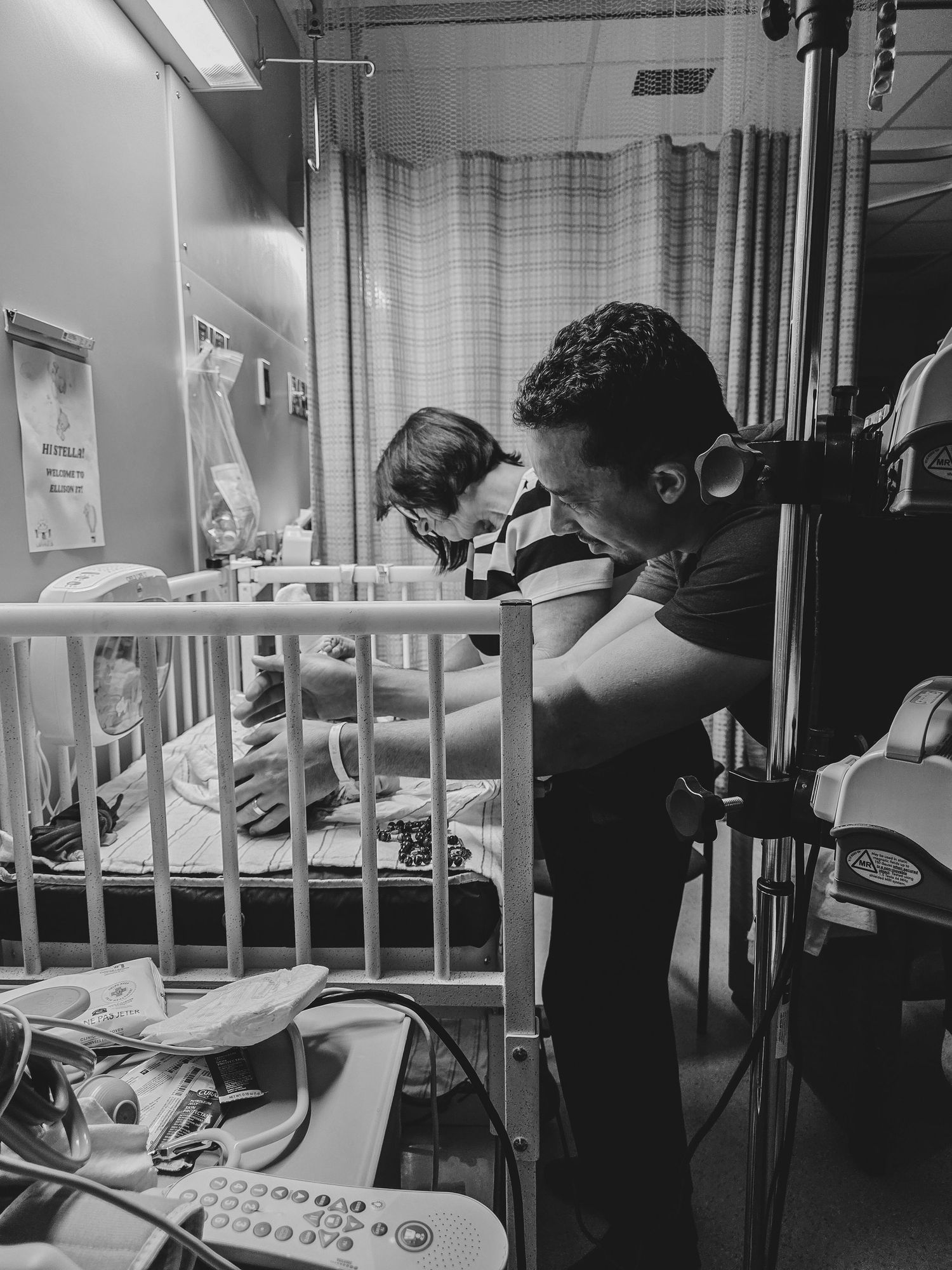 Two parents lean over a hospital crib in a pediatric room, tenderly caring for their baby beside medical monitors in black and white.