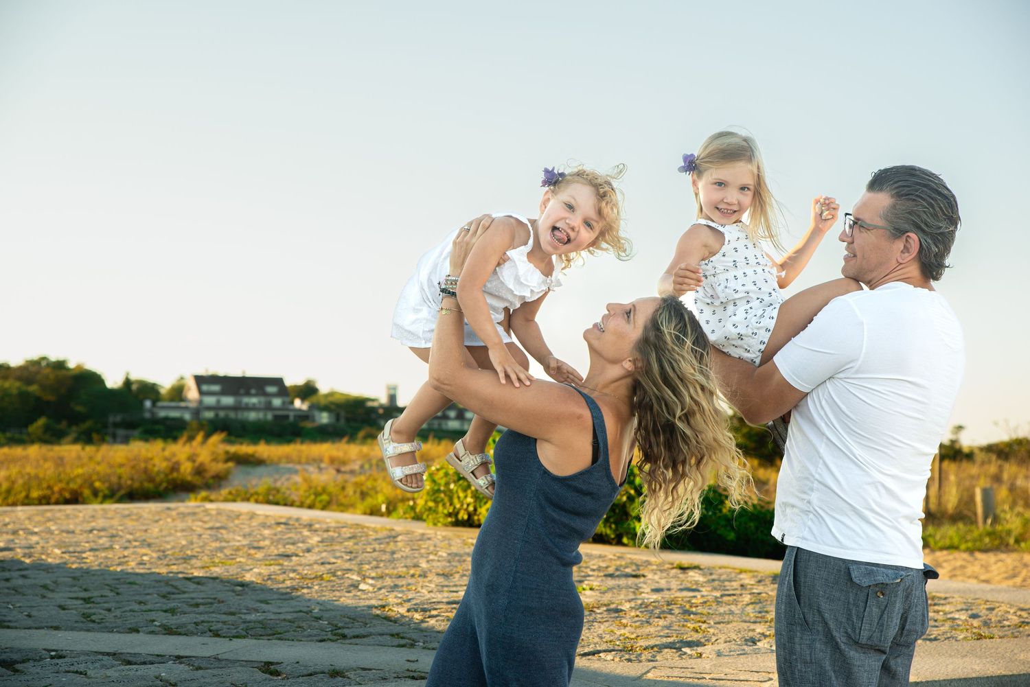 Happy family with two toddler girls being lifted by parents on a sunny beach path.