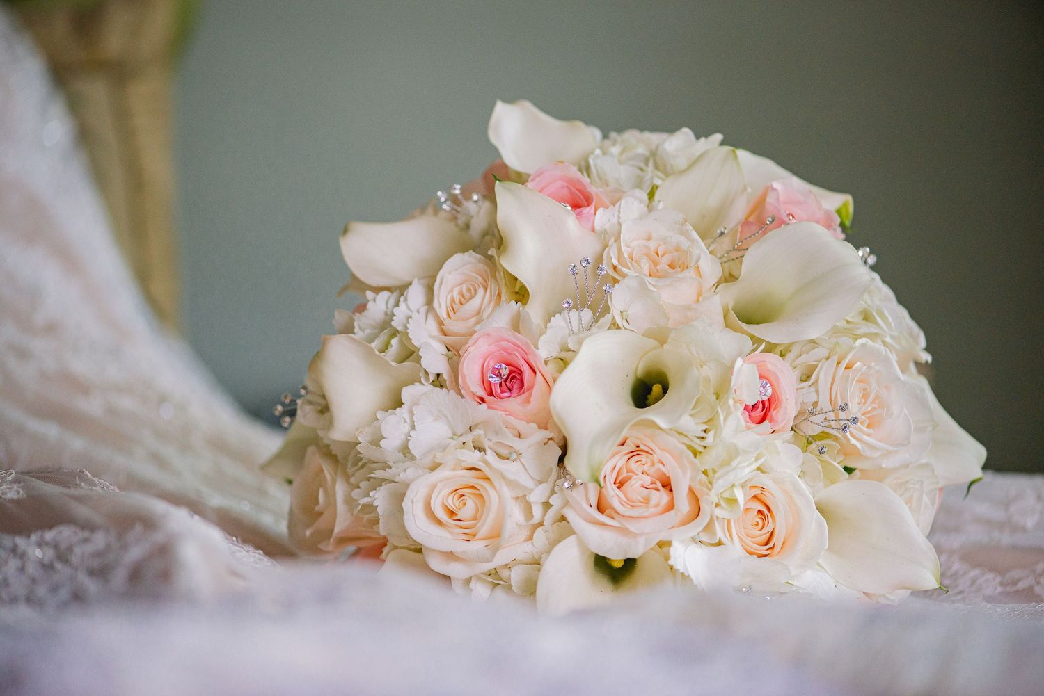 Elegant bridal bouquet of white calla lilies and blush pink roses resting on lace wedding dress fabric.
